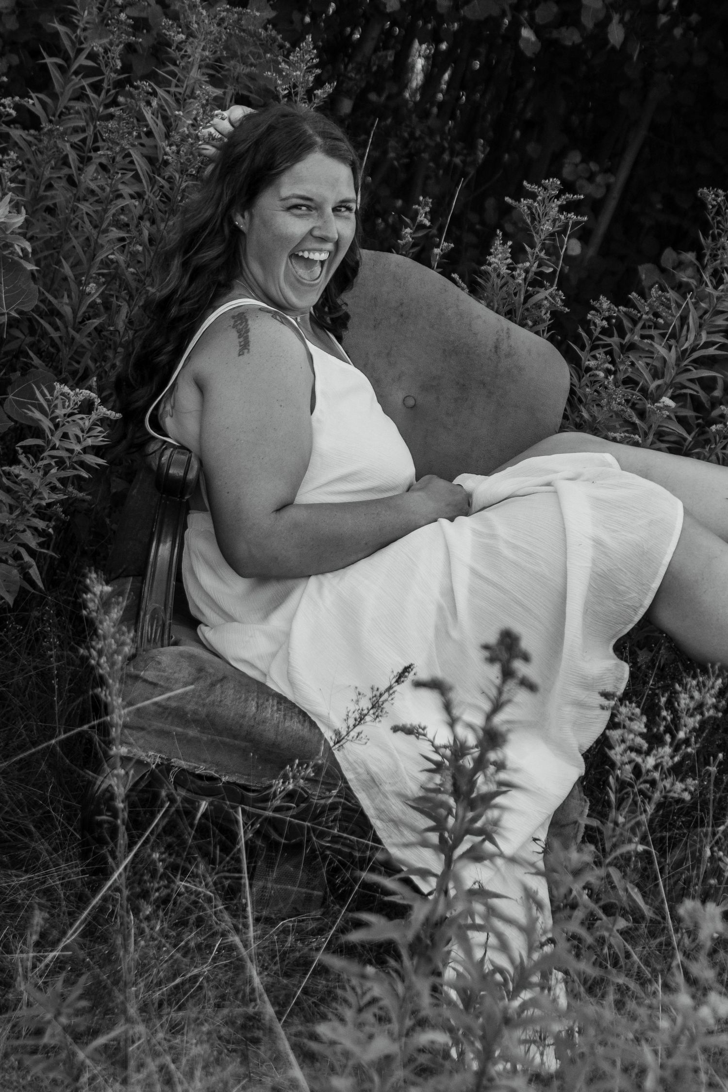 Black and white portrait outdoors showing someone relaxing in a white dress on a garden chair amid foliage.