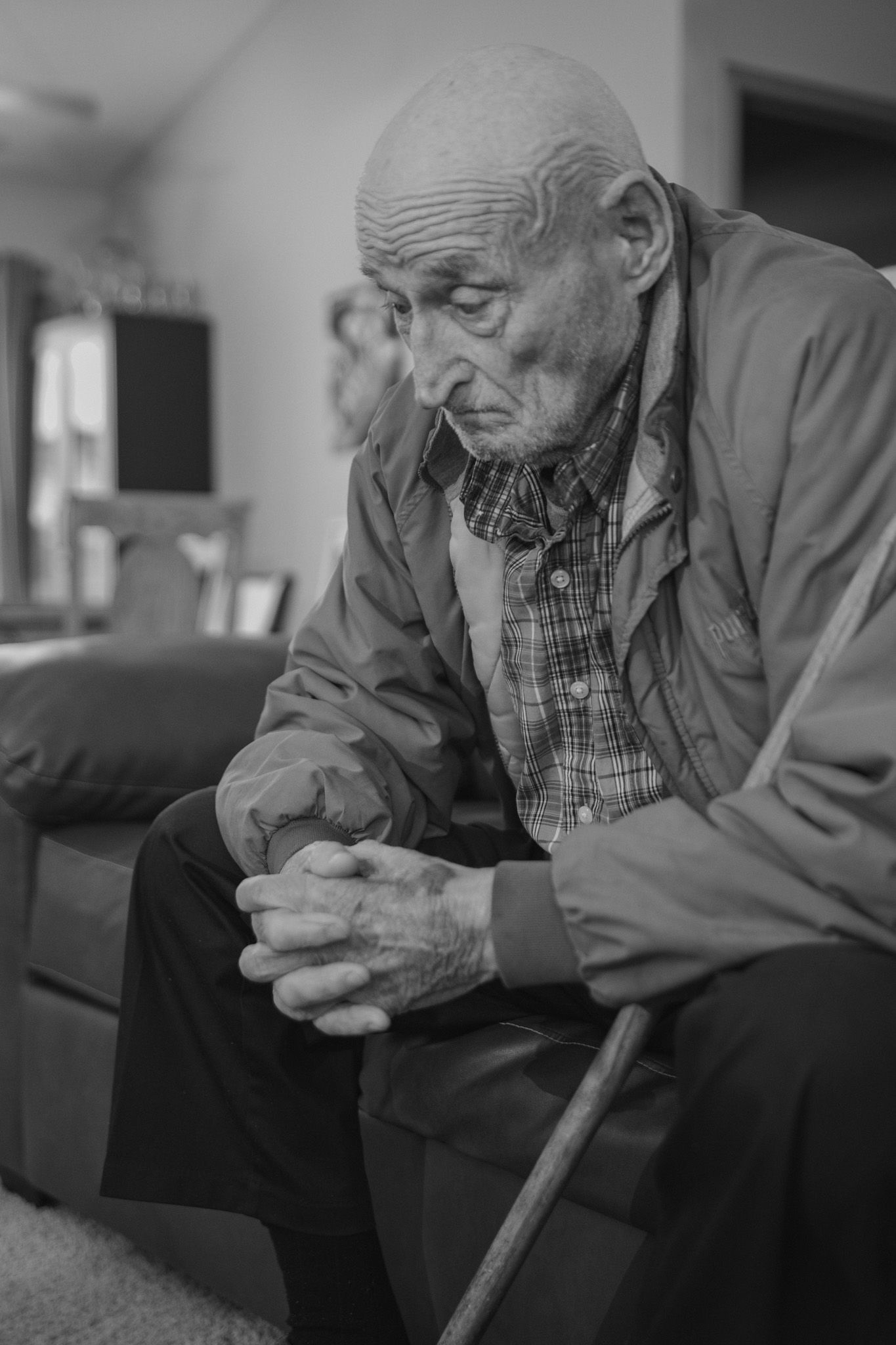 Elderly man with cane sits pensively, hands clasped, in black and white portrait photo.