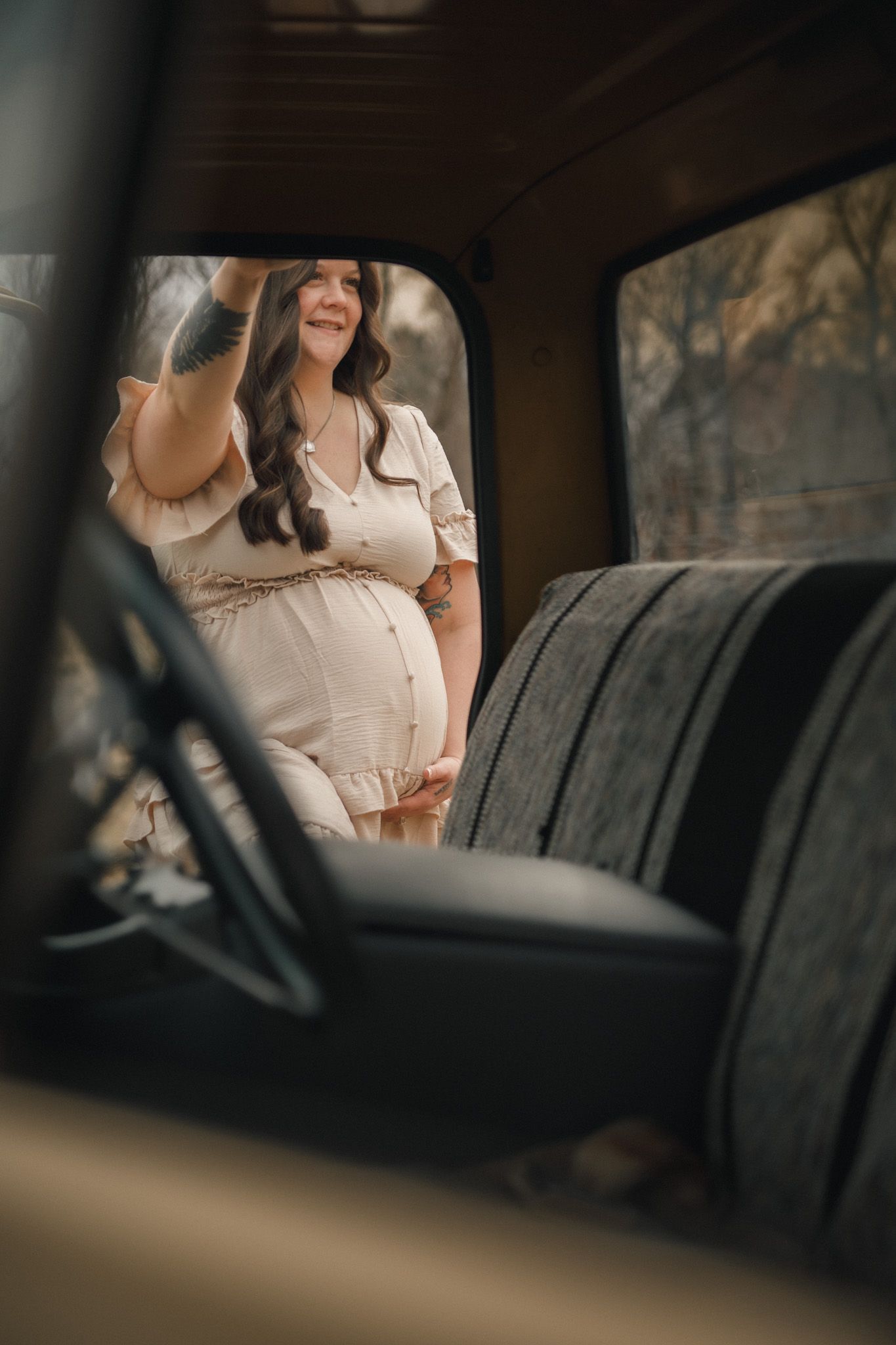 Pregnant woman in cream dress viewed through vintage truck window during outdoor rustic maternity photoshoot.