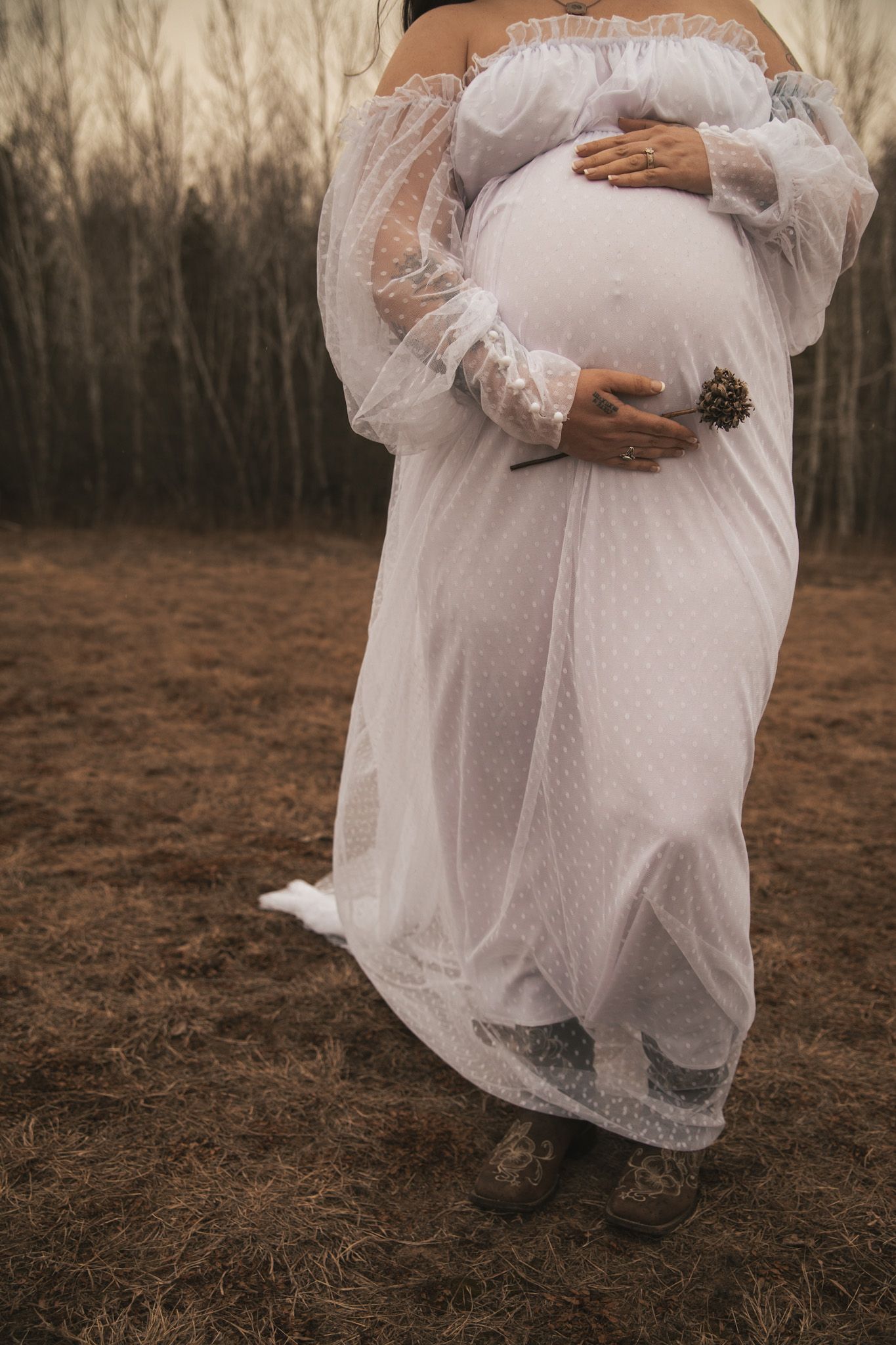 Close-up of pregnant woman in white lace dress cradling baby bump outdoors.
