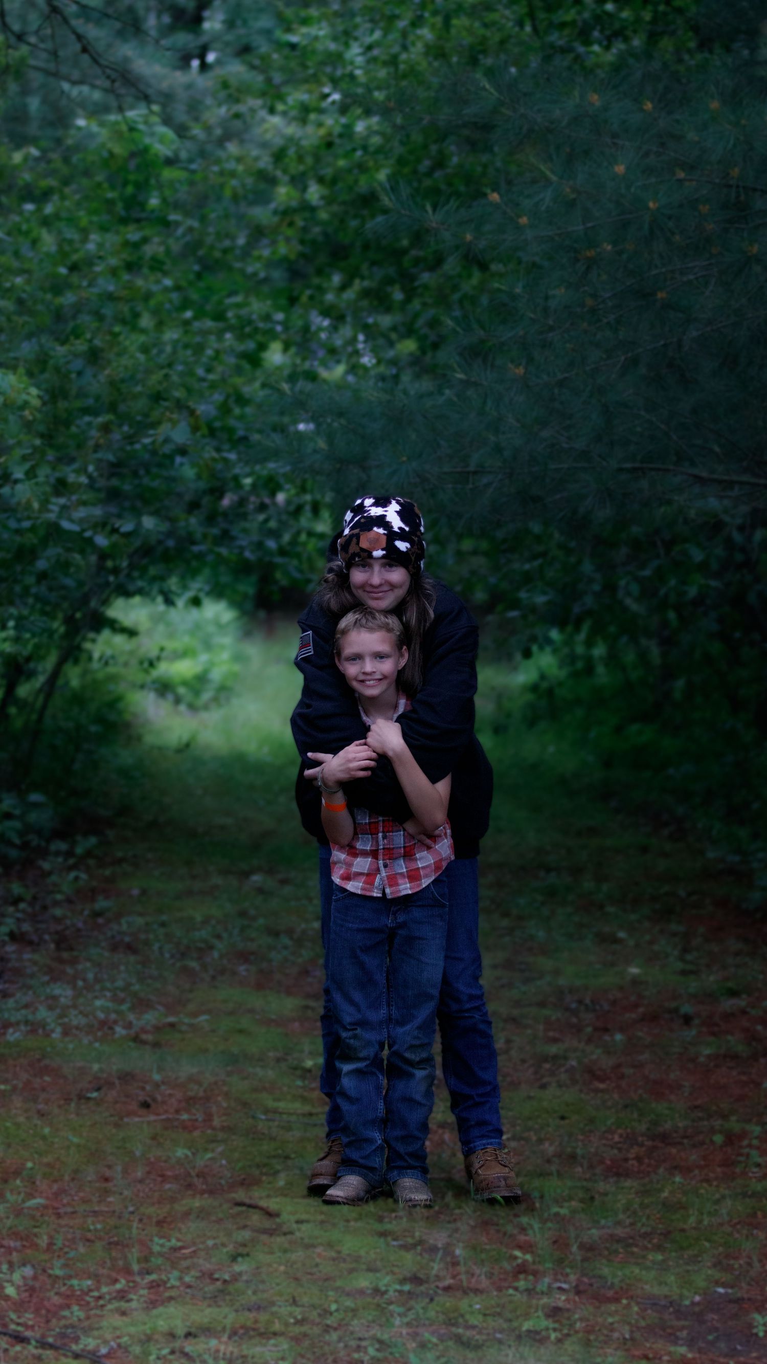 Two people standing together in a wooded area on a dim forest path wearing casual clothing.