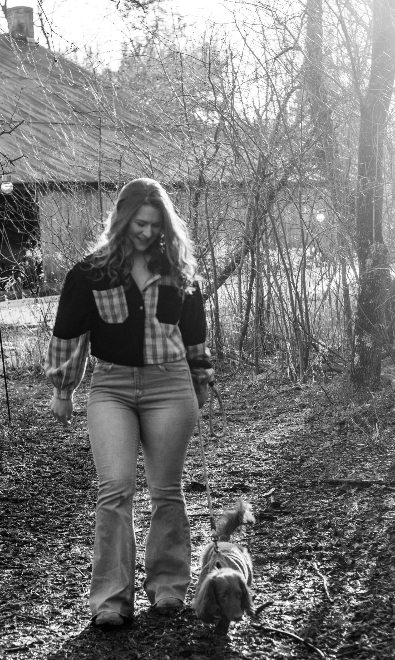 Woman in plaid shirt and flare jeans smiling while walking a small dog on a rural path in black and white.