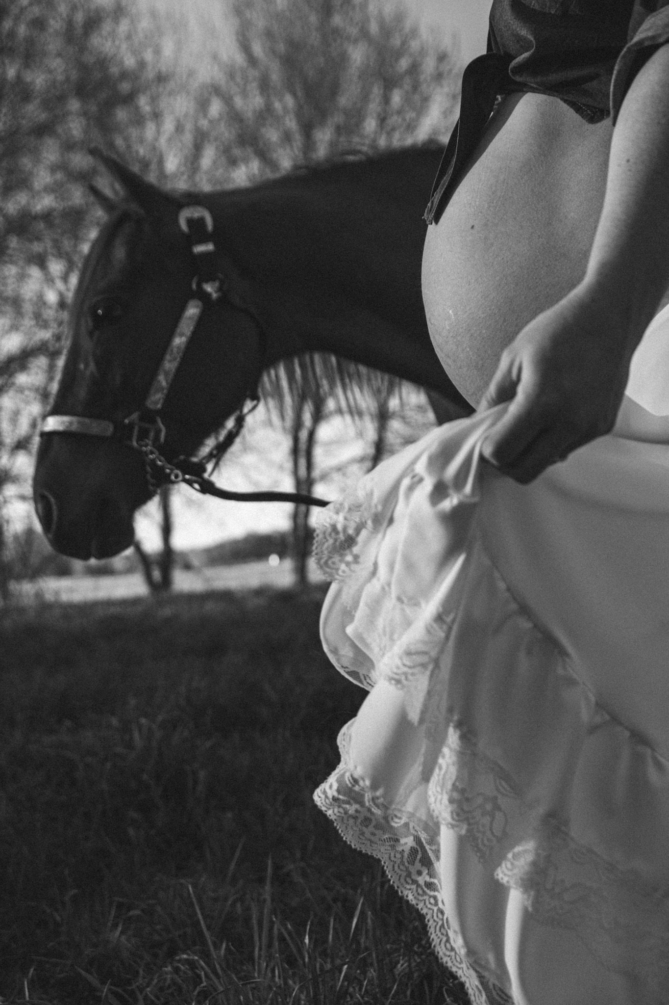 Black and white maternity photo of pregnant woman in lace dress standing beside a horse outdoors.