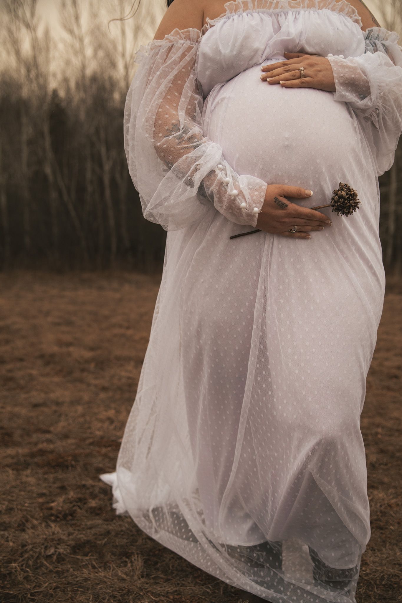 Close-up of pregnant woman cradling baby bump in sheer white off-shoulder maternity gown outdoors.