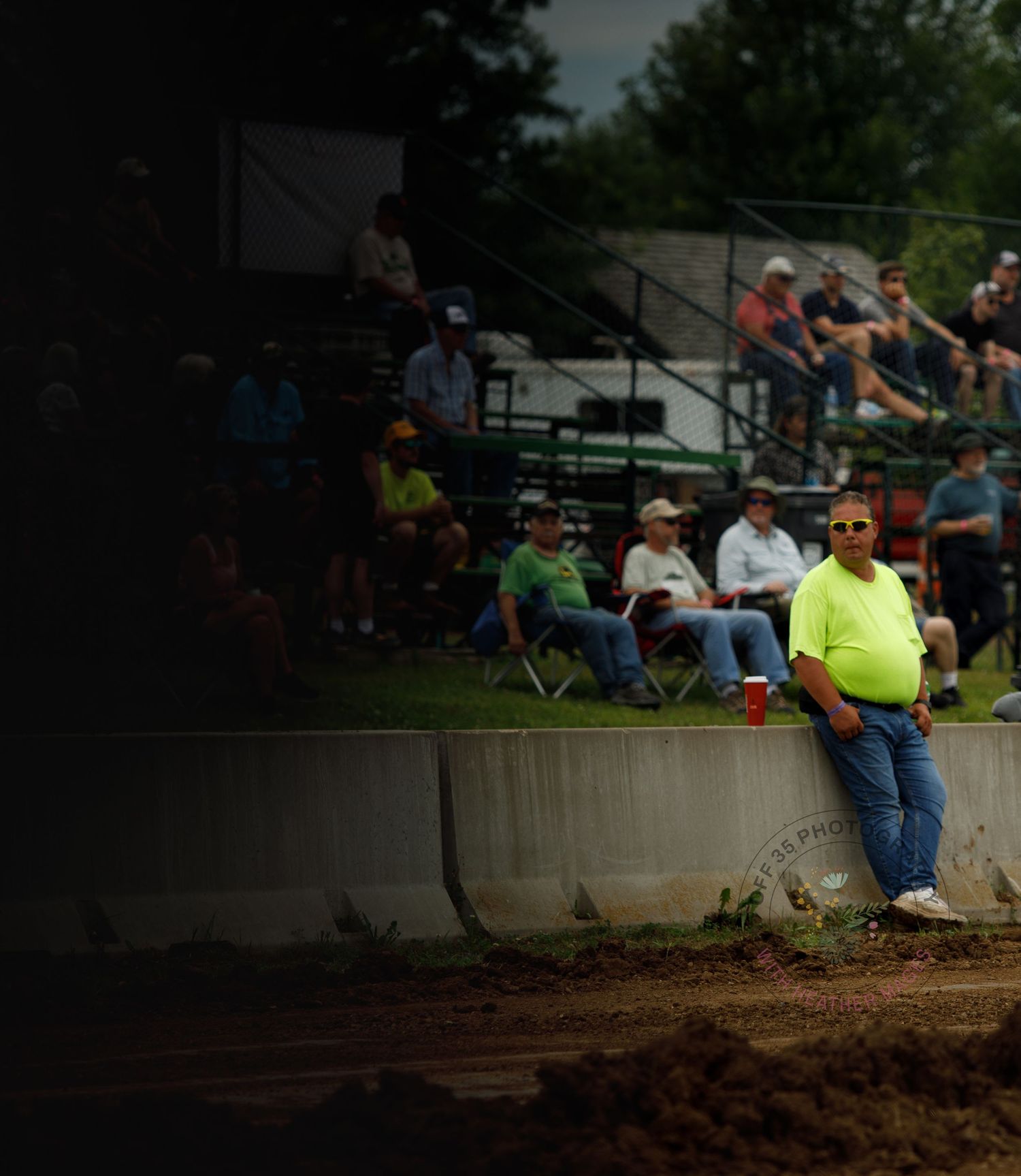 Spectators gather around a dirt track watching an outdoor event from folding chairs and concrete barriers.