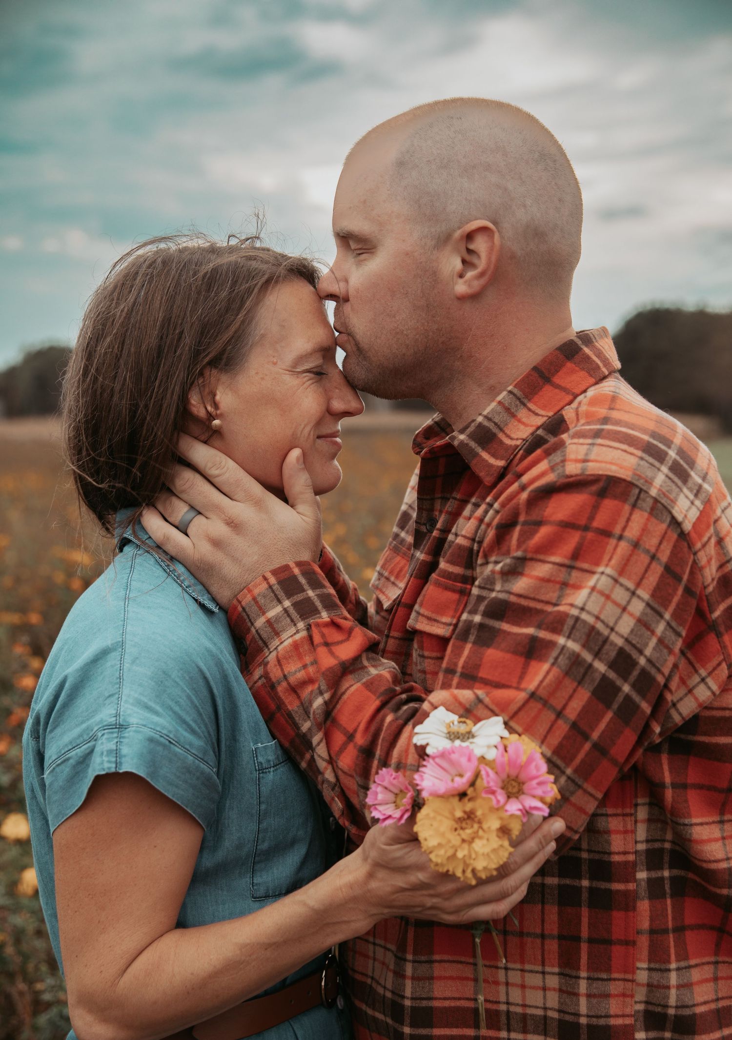 A couple shares an intimate moment in a field while holding flowers against a moody sky background.