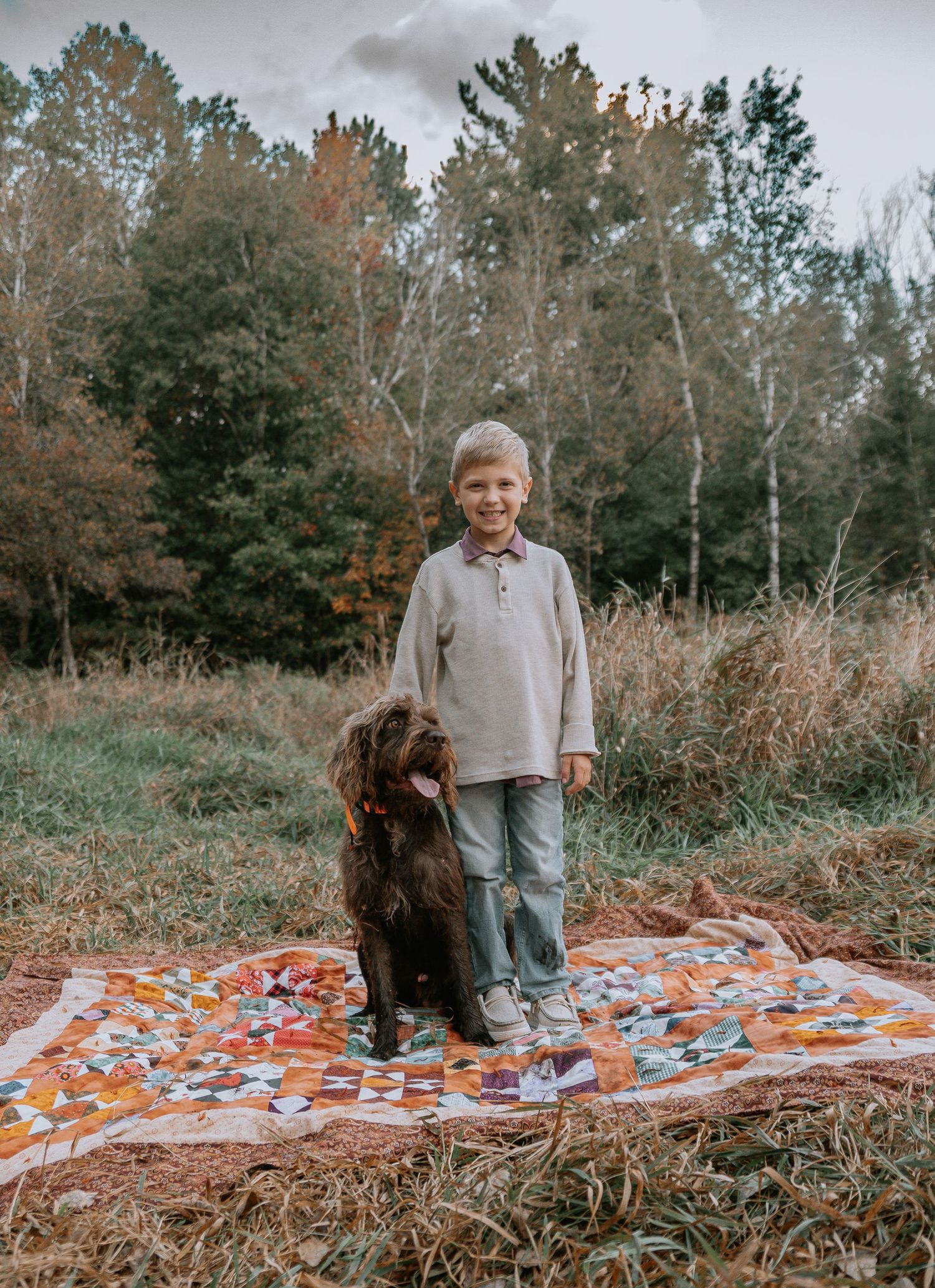 A person and a brown dog sit together on a patterned blanket in a wooded autumn setting.