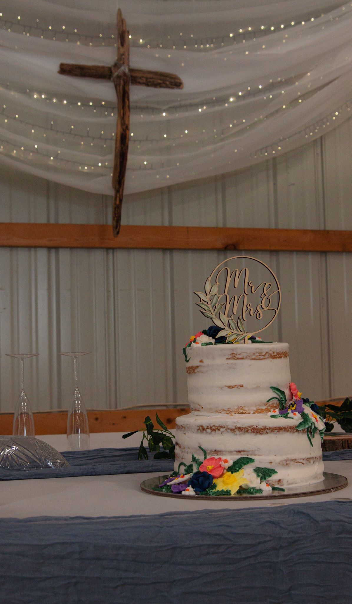 A semi-naked wedding cake with floral decorations and a Mr & Mrs topper displayed against draped fabric with string lights.
