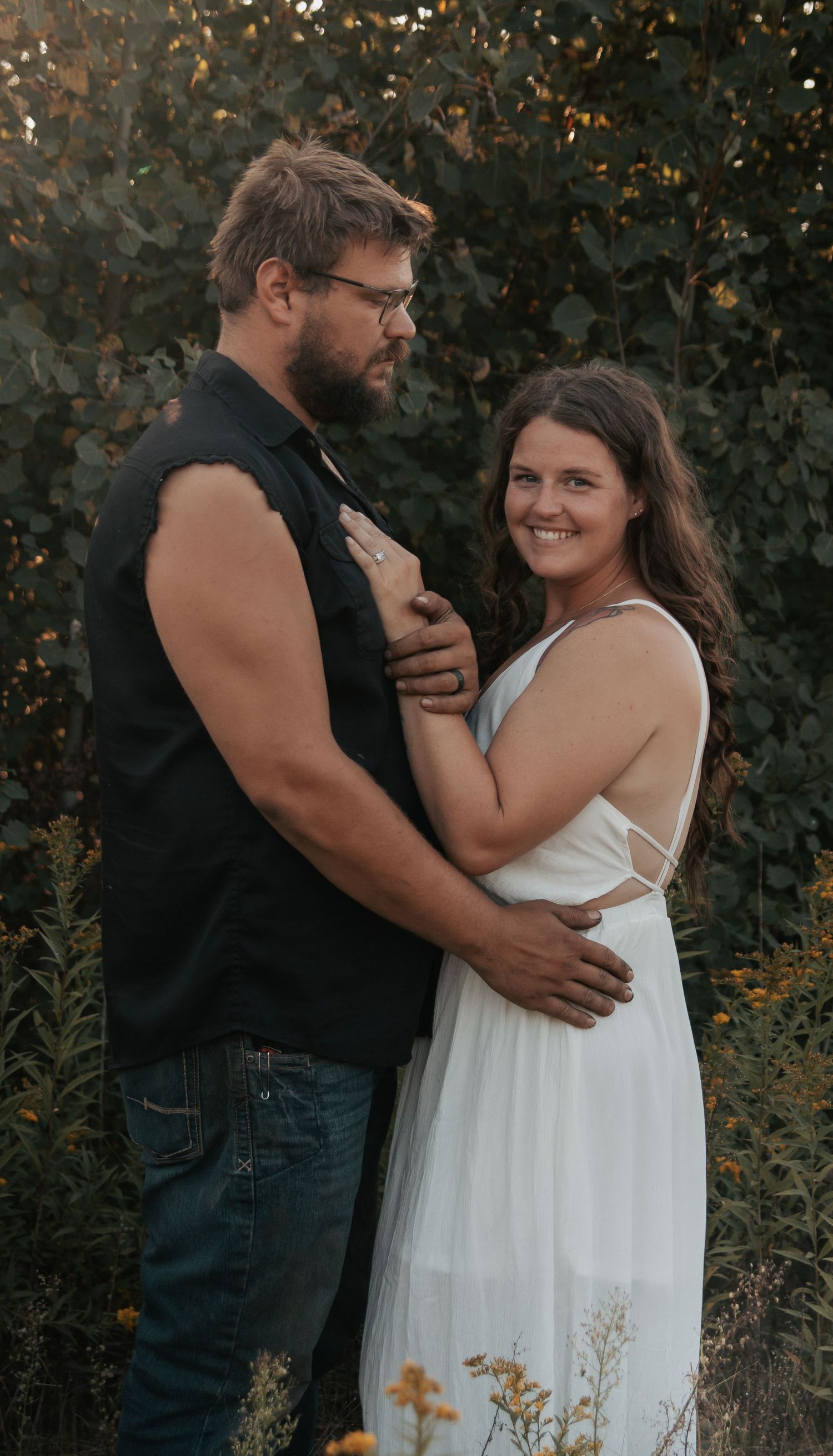 A romantic couple embraces outdoors in a garden setting with wildflowers at sunset.