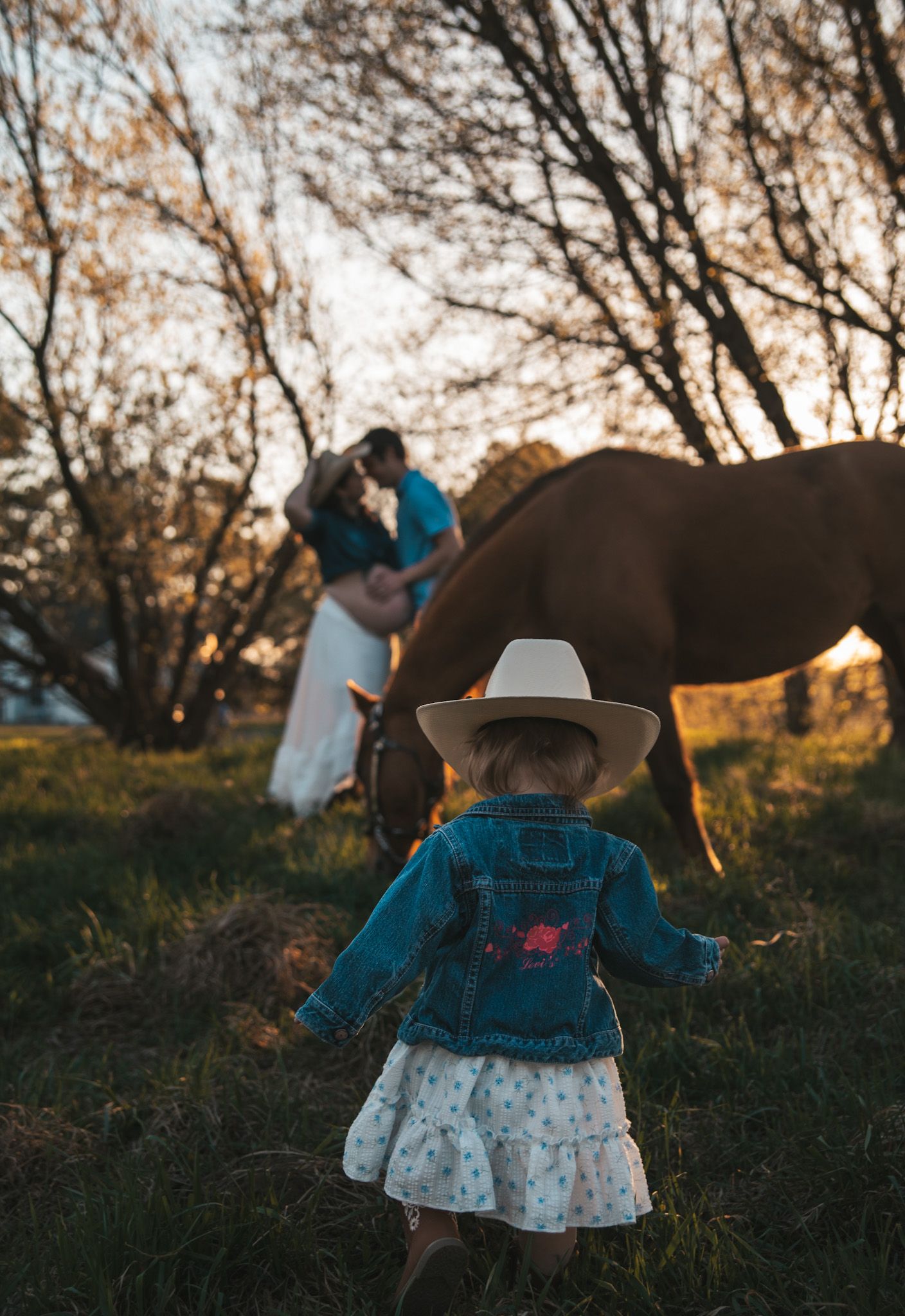 Young girl in denim jacket and white cowboy hat walks toward couple and horse at sunset.