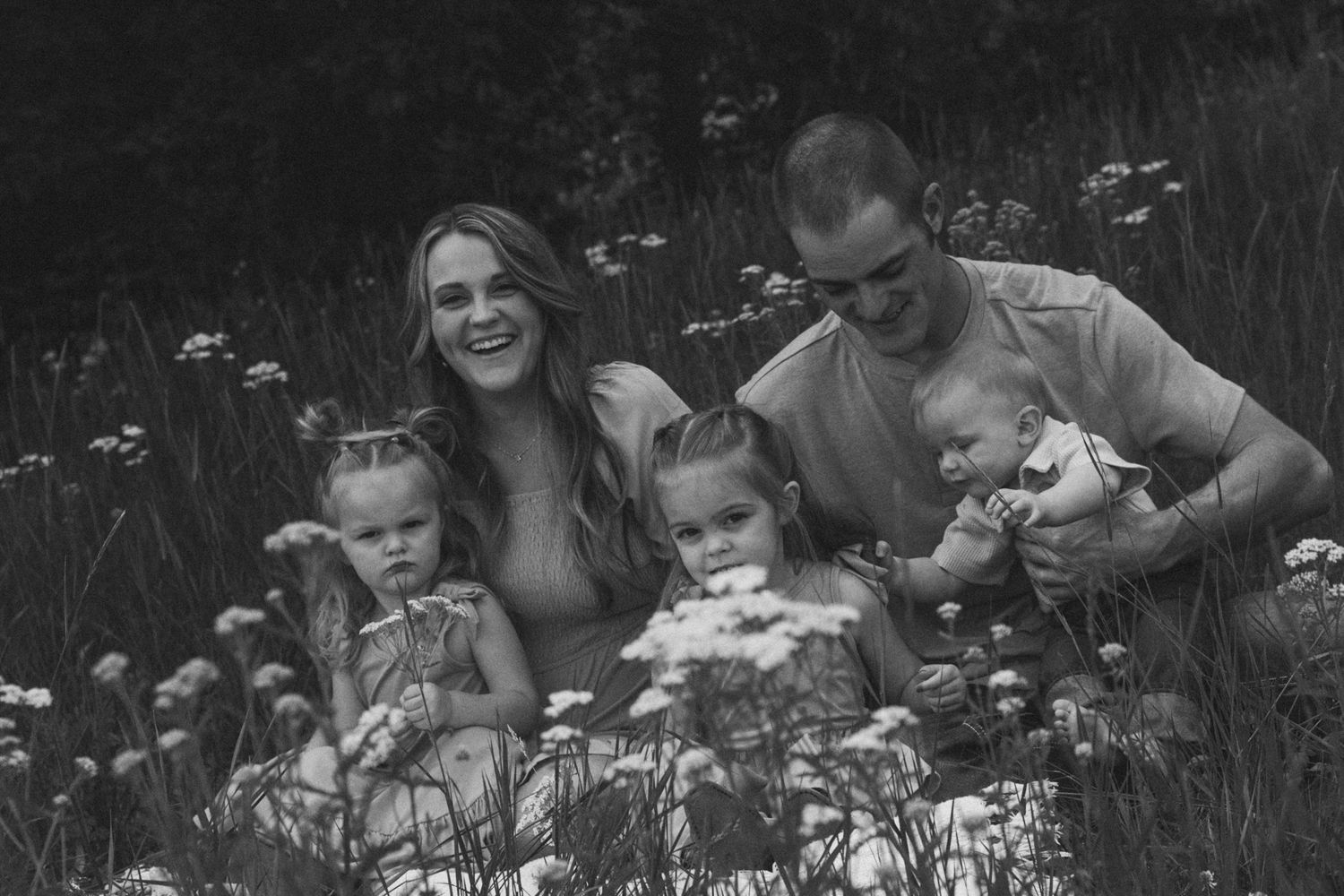 Black and white family photo of parents and three young children sitting in a wildflower meadow, smiling joyfully.