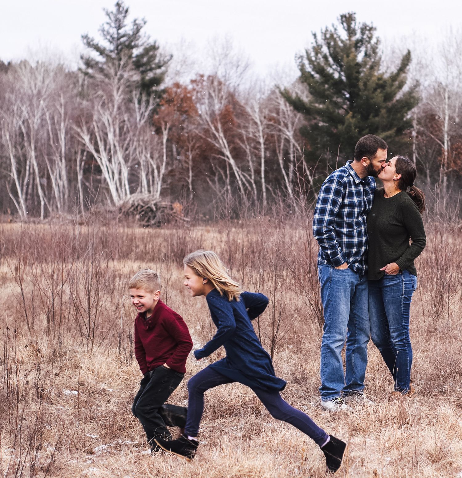 A couple walks together through a field of dried grass and winter trees, engaging in playful poses and interactions.