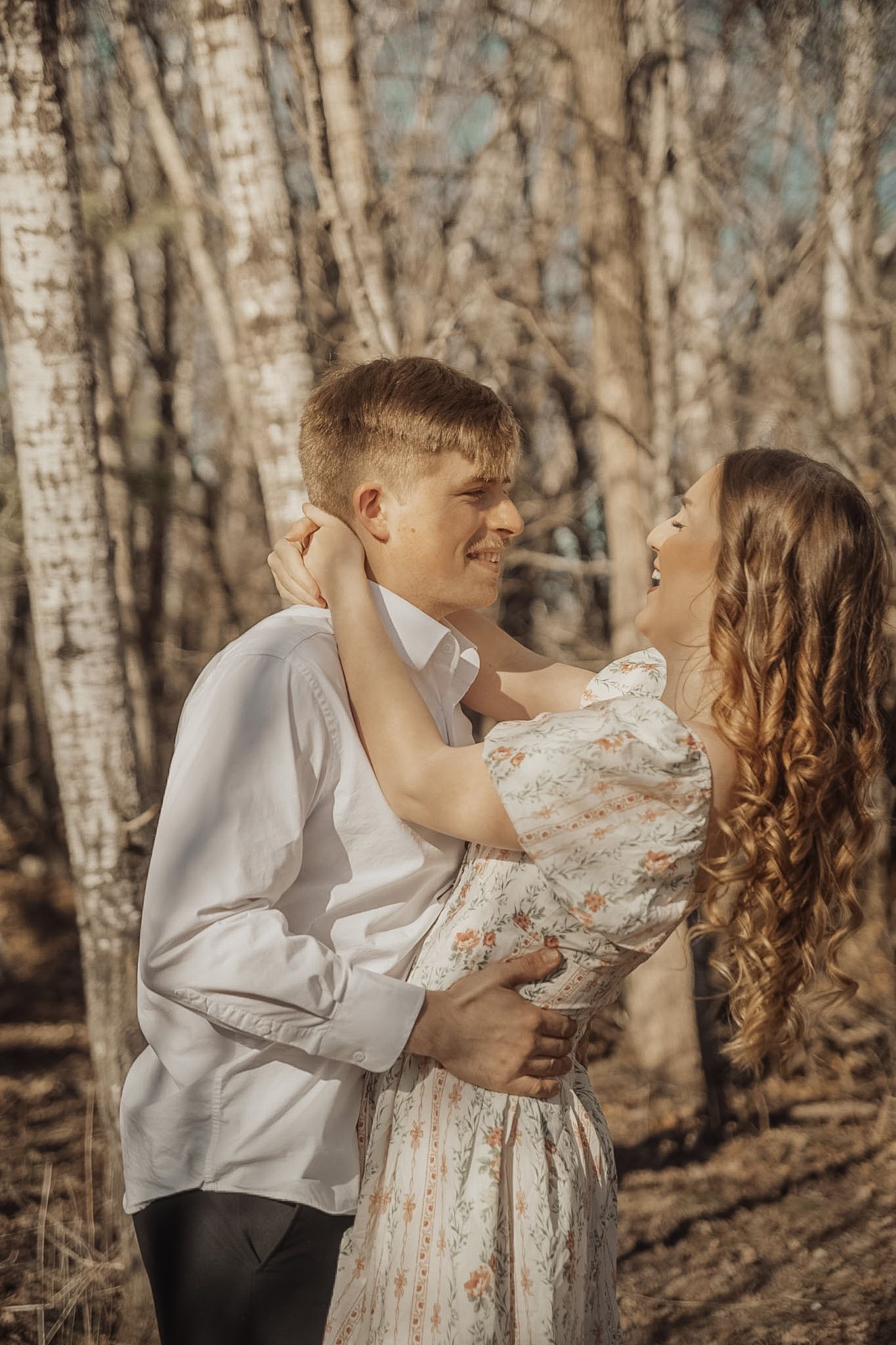 A young couple embracing and laughing together in a sunny birch forest, woman in floral dress, man in white shirt.