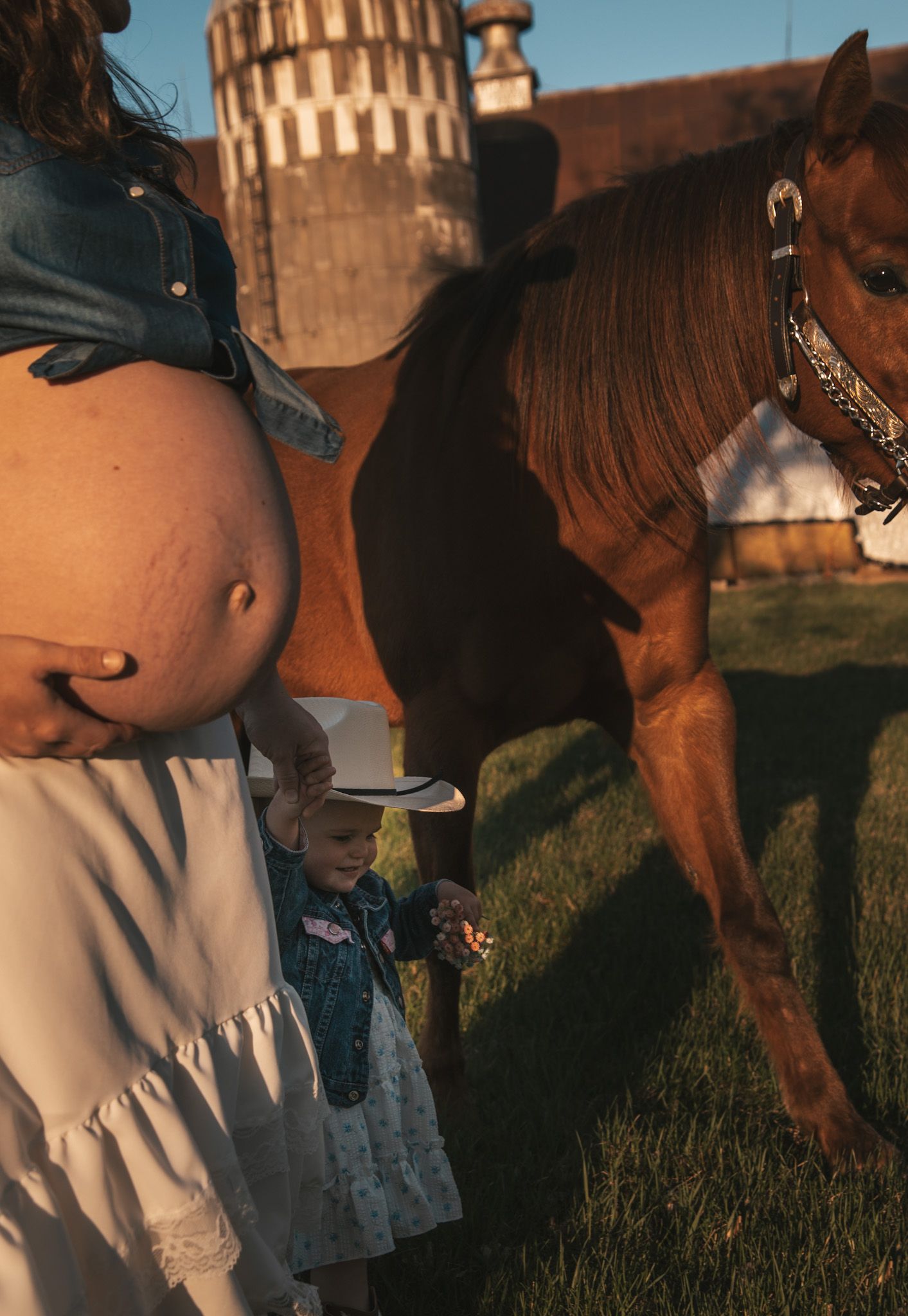 Pregnant woman with toddler in cowboy hat standing beside a chestnut horse near a red barn at golden hour.