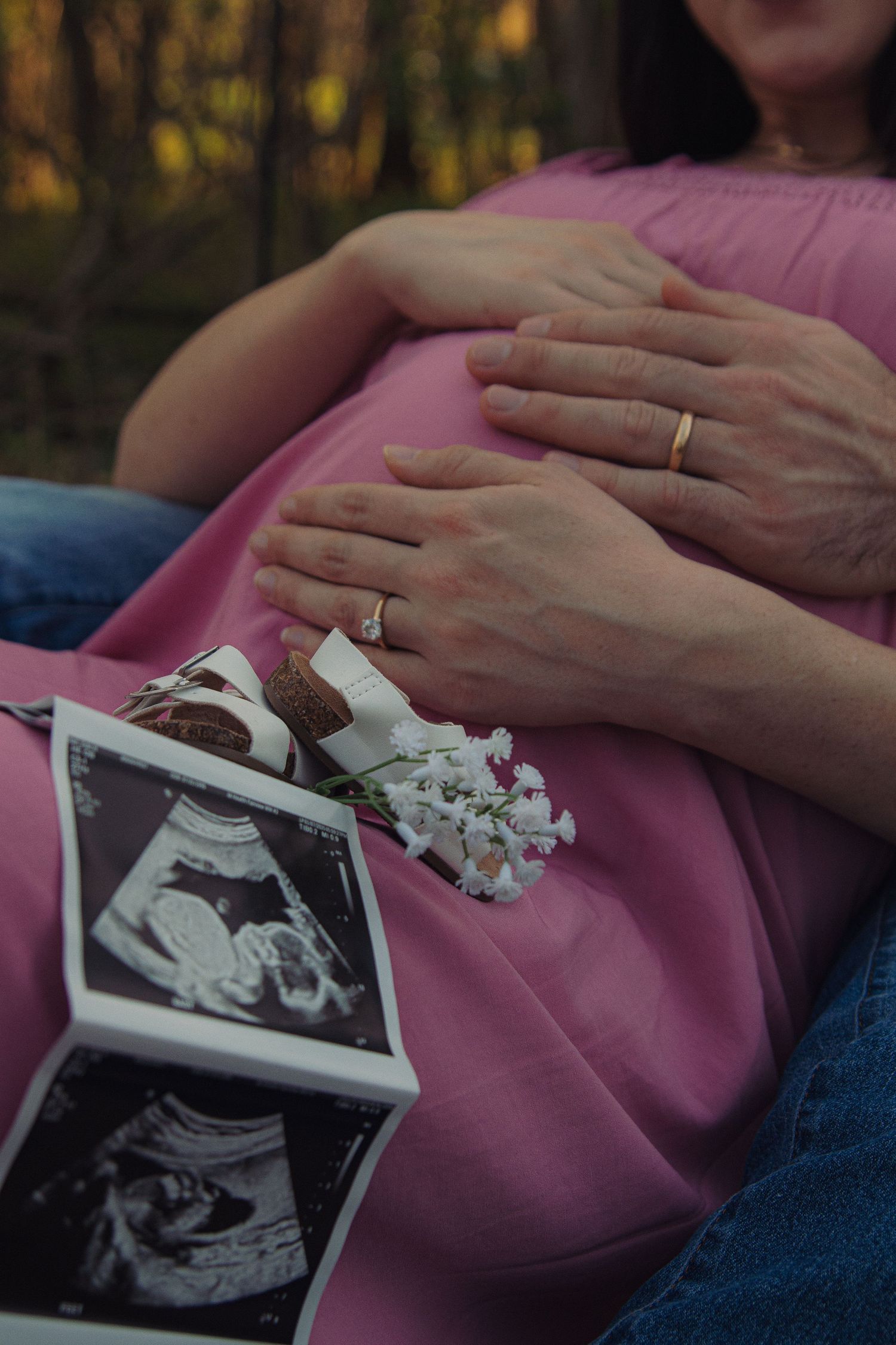 Hands rest on a pink dress with ultrasound photos and dried flowers, showing wedding rings in warm evening light.