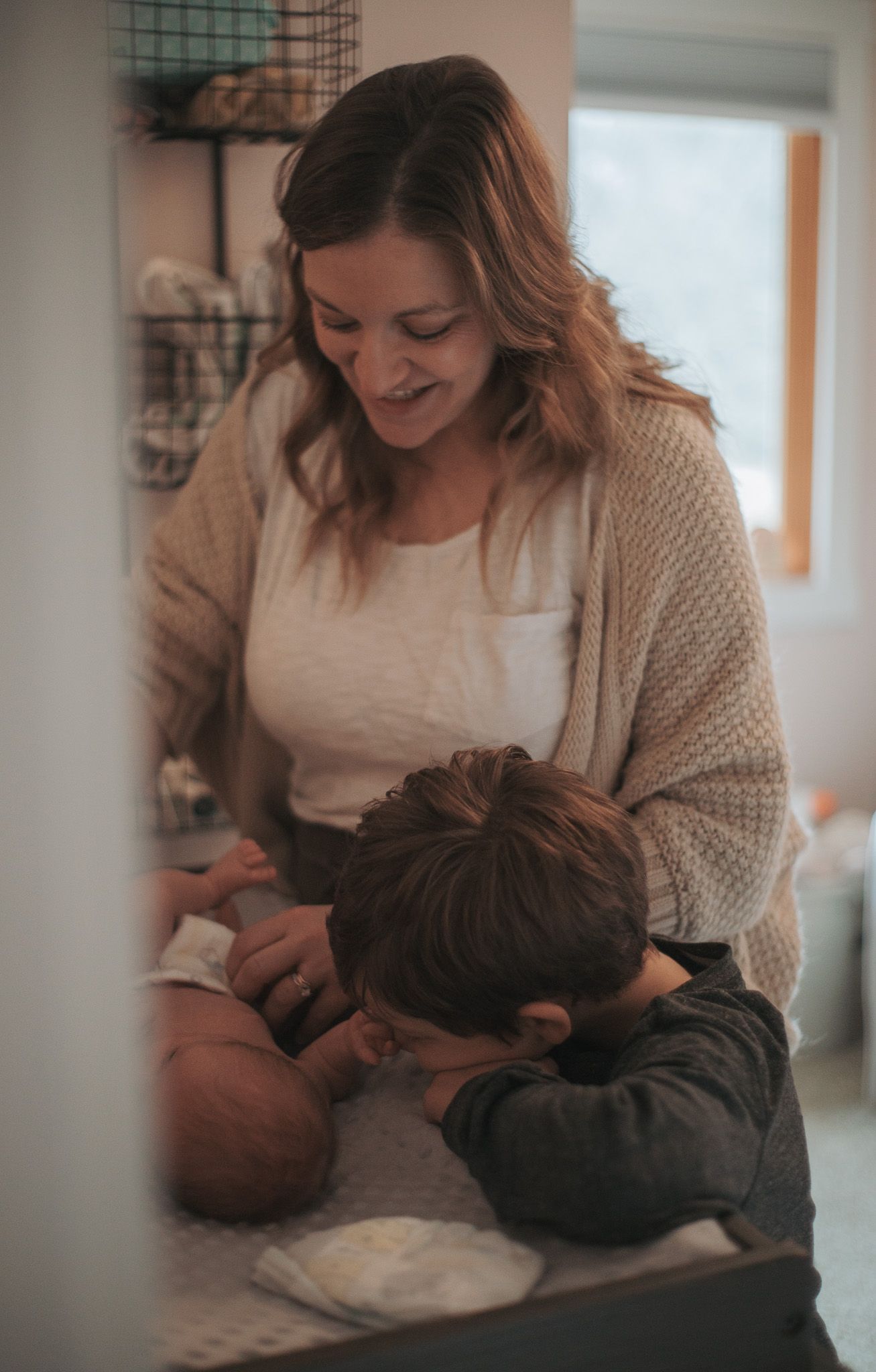 A smiling mother watches as a young child leans over to meet their newborn sibling on a changing table.