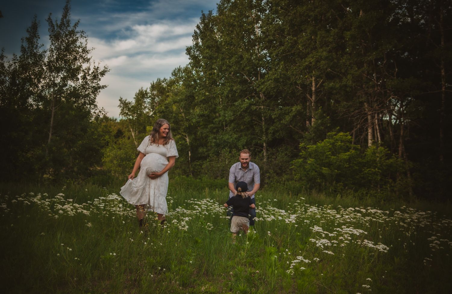 Pregnant woman in white dress with partner and dog in a lush green meadow surrounded by trees during a maternity photoshoot.