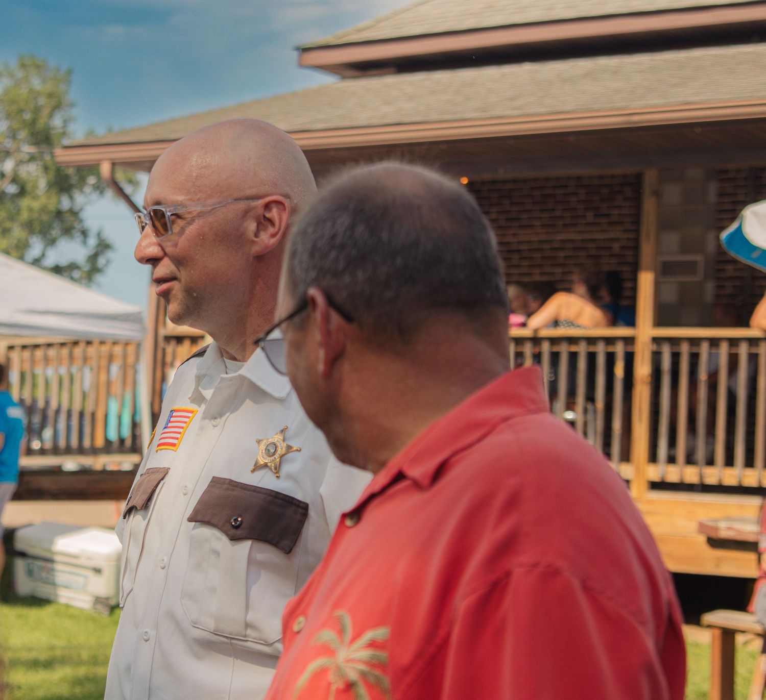 A uniformed sheriff deputy and person in red shirt converse outside a building with a wooden porch.