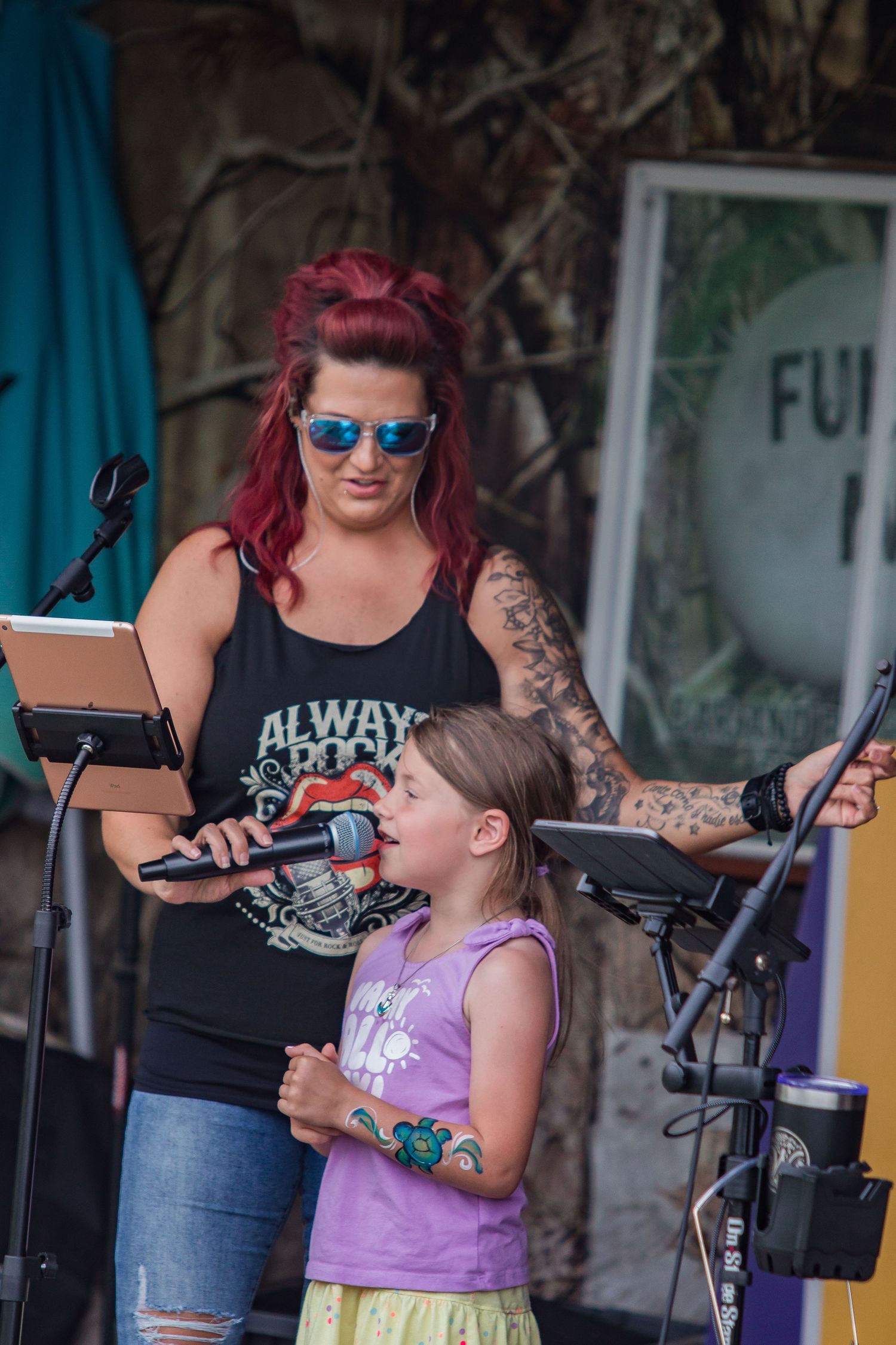 A performer and young girl share a microphone on an outdoor stage during a music event.