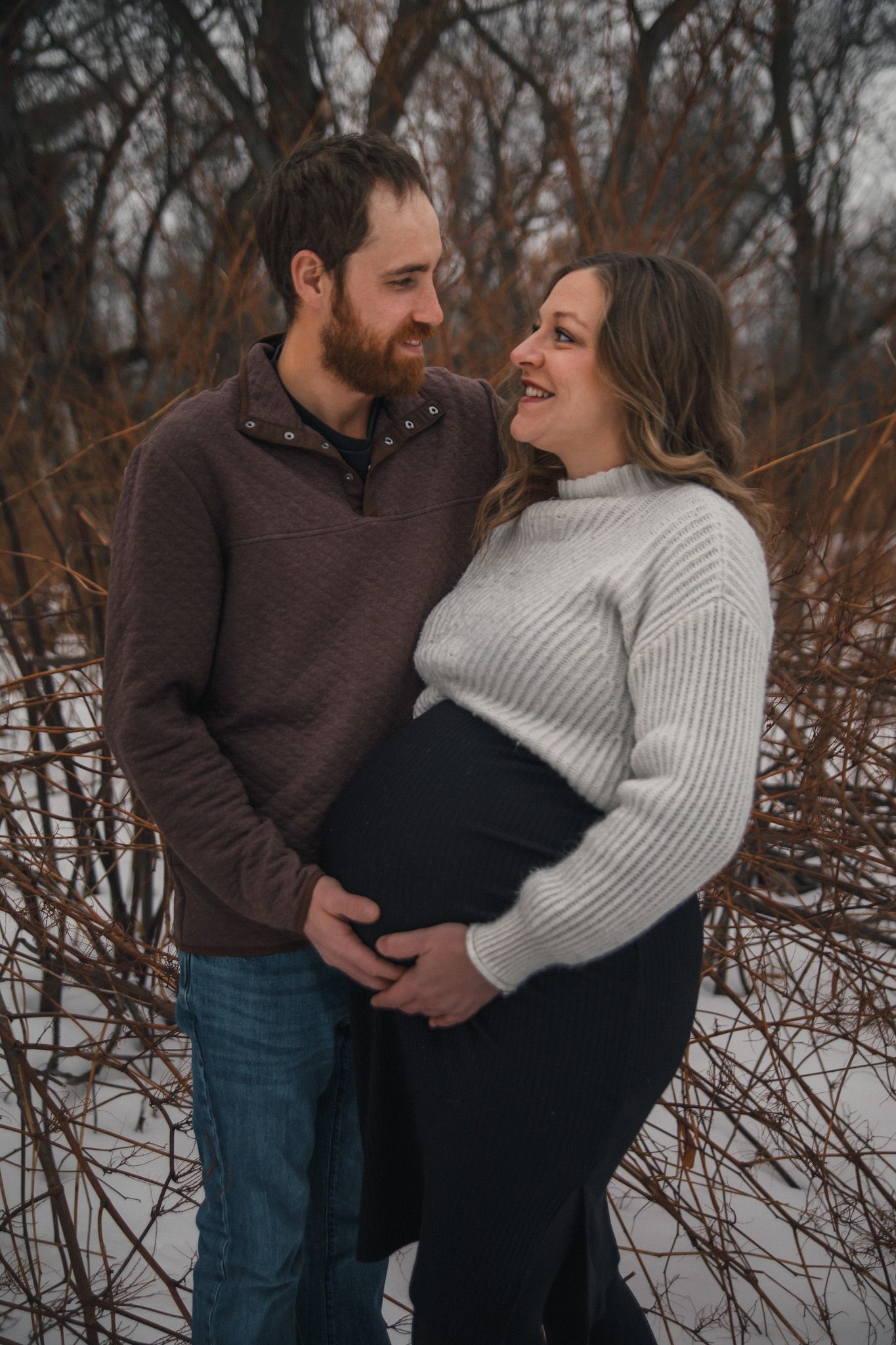 A couple shares an intimate moment in a snowy winter setting, wearing cozy sweaters among bare branches.