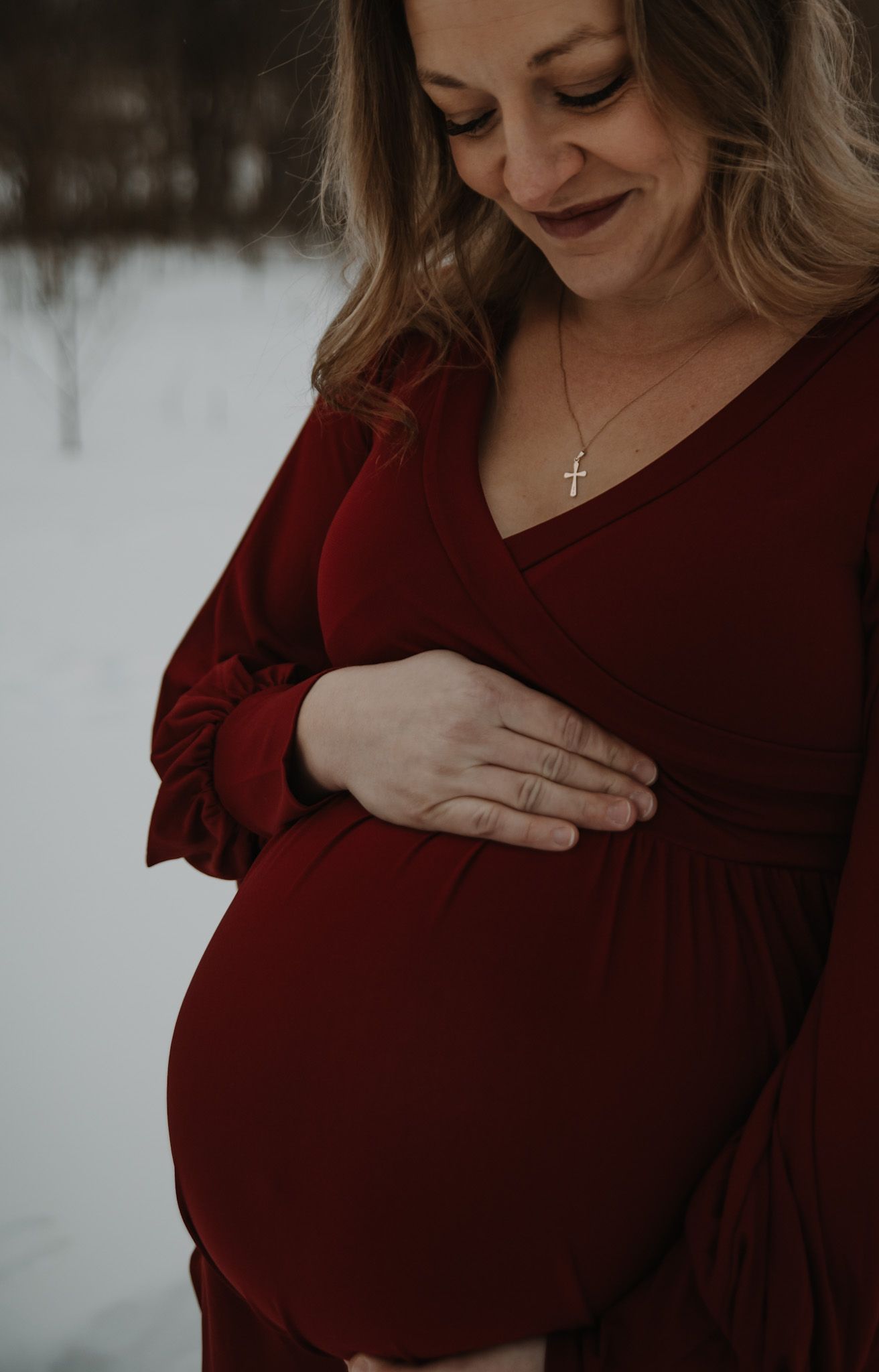 Close-up shot of pregnant woman in burgundy dress tenderly holding her baby bump against snowy background.