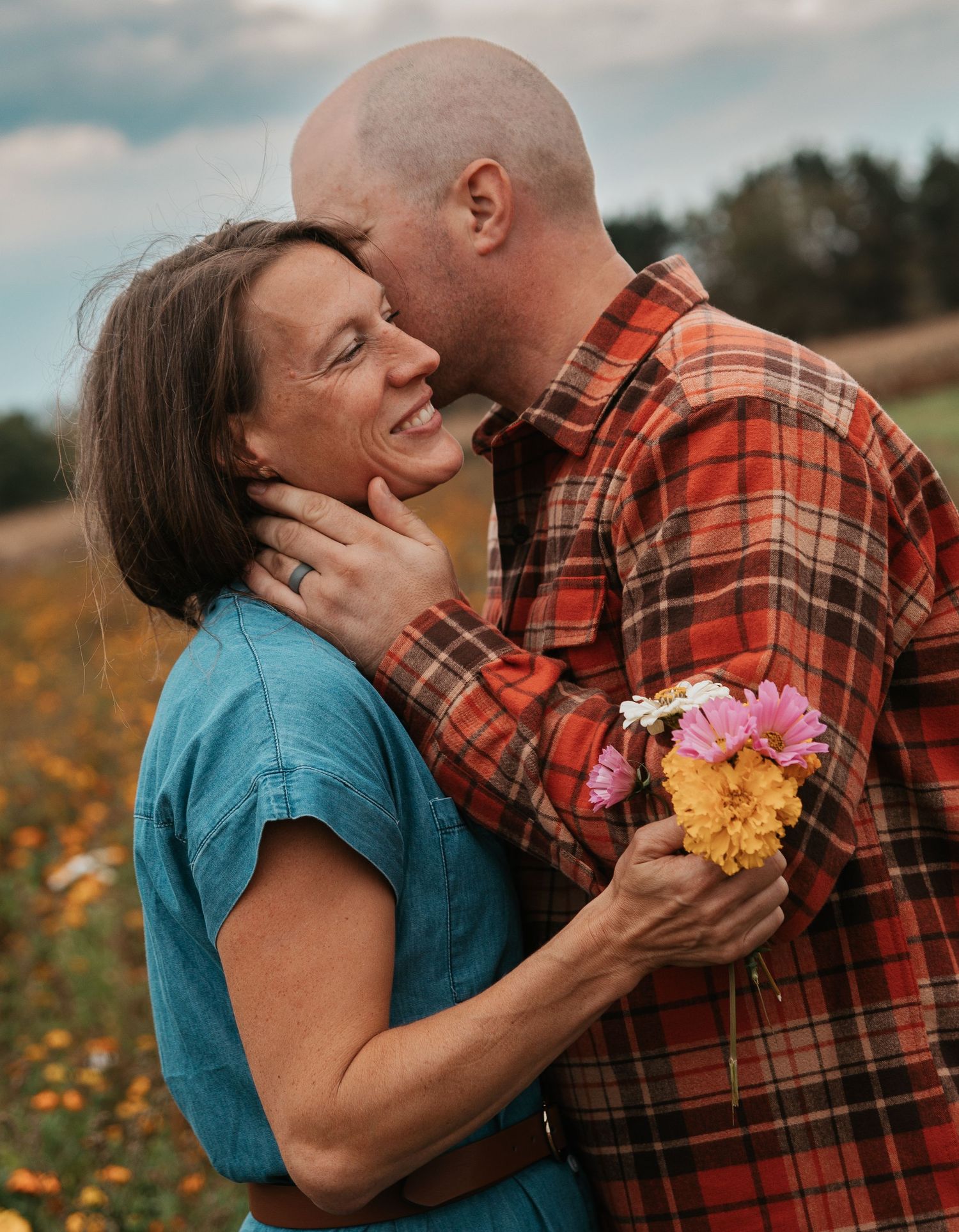 A couple shares an intimate moment in a field of yellow wildflowers, with the person in blue holding a bouquet.