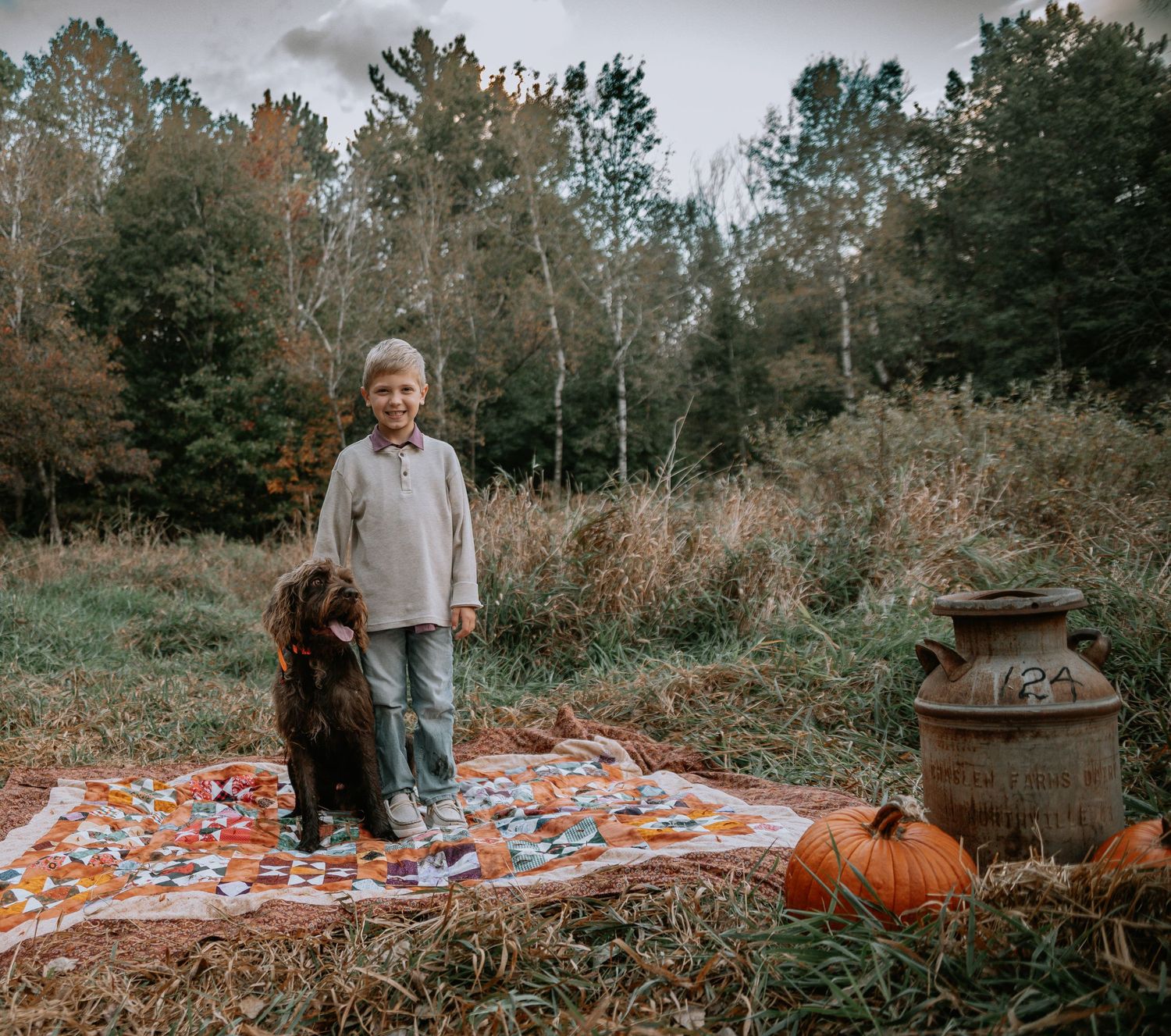A couple and their dog stand on a cozy fall blanket surrounded by autumn foliage and pumpkins in a forest setting.