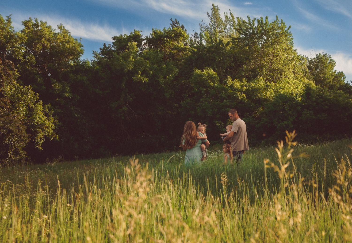 Family of three standing in a lush green meadow surrounded by tall trees on a sunny day.