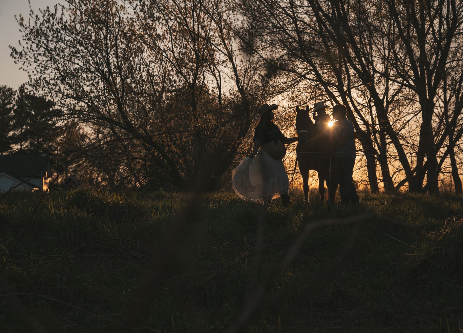Silhouette of couple and horse against golden sunset light through bare trees at dusk.
