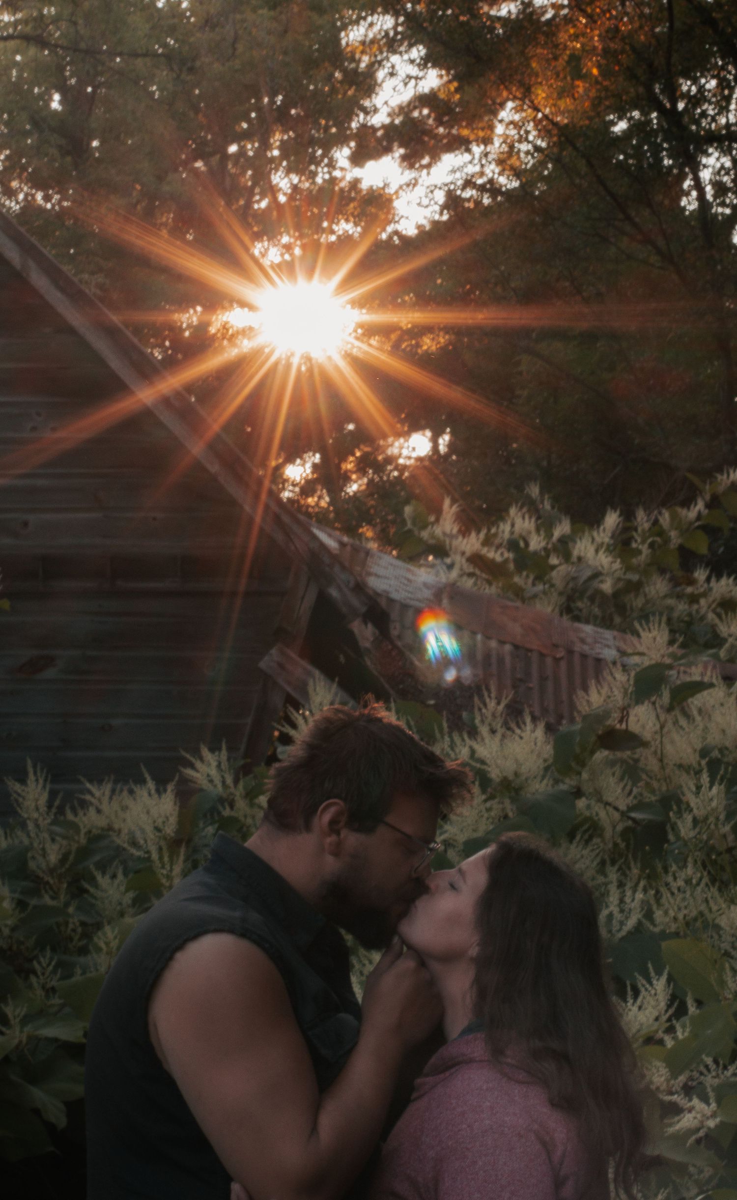A couple embraces in a romantic kiss against a sunset backdrop with sunbeams streaming through trees and white flowers.