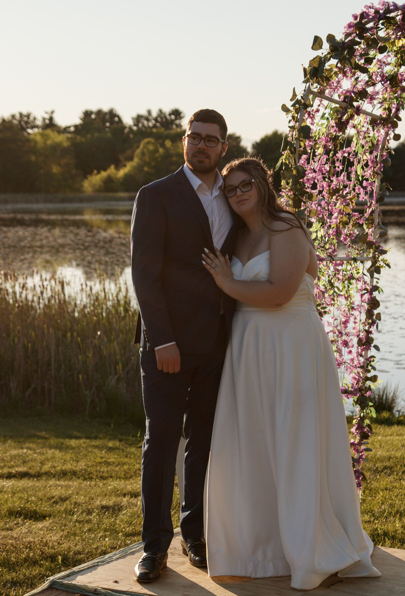 Couple embraces tenderly by a lake during golden hour beneath blooming cherry blossoms on a spring evening.