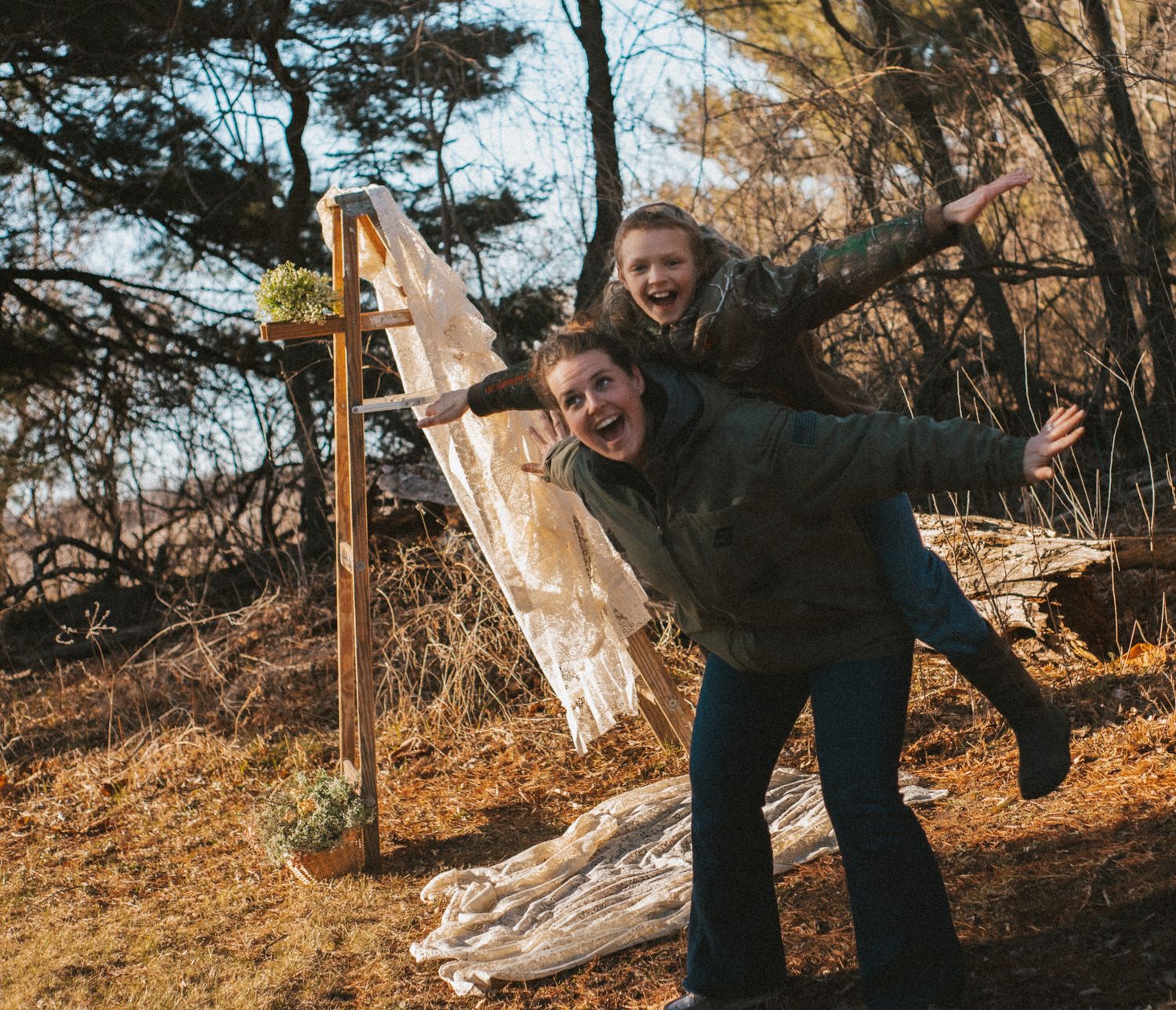 Two laughing women pose playfully outdoors in a wooded area, one giving the other a piggyback ride near a rustic arch.