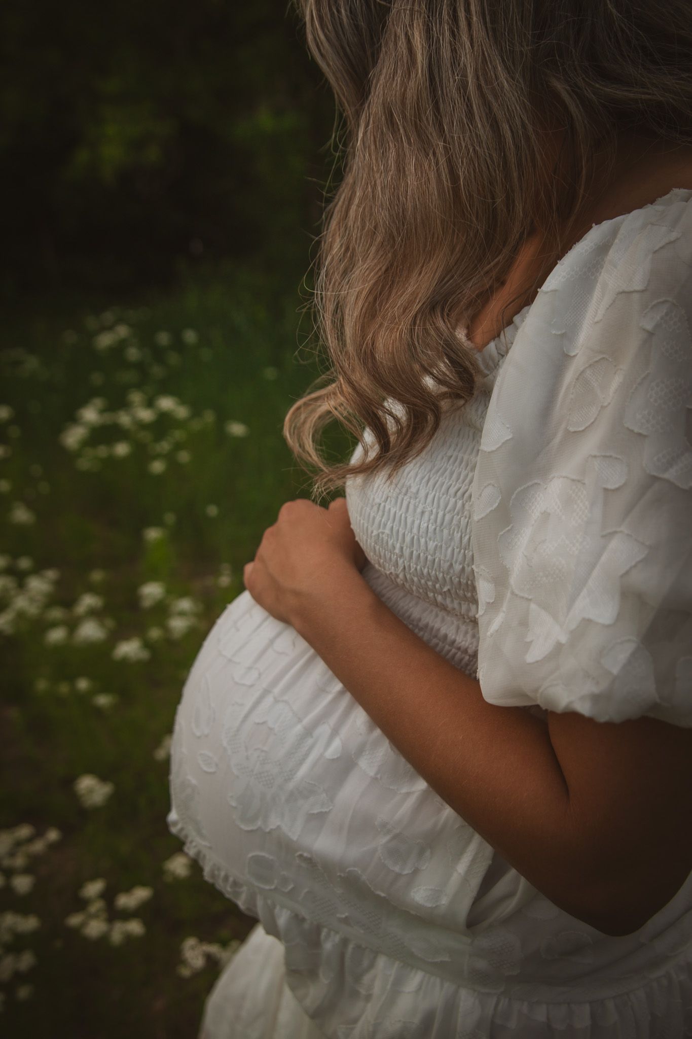 Pregnant woman in white ruffled dress cradling baby bump outdoors in a field of wildflowers.