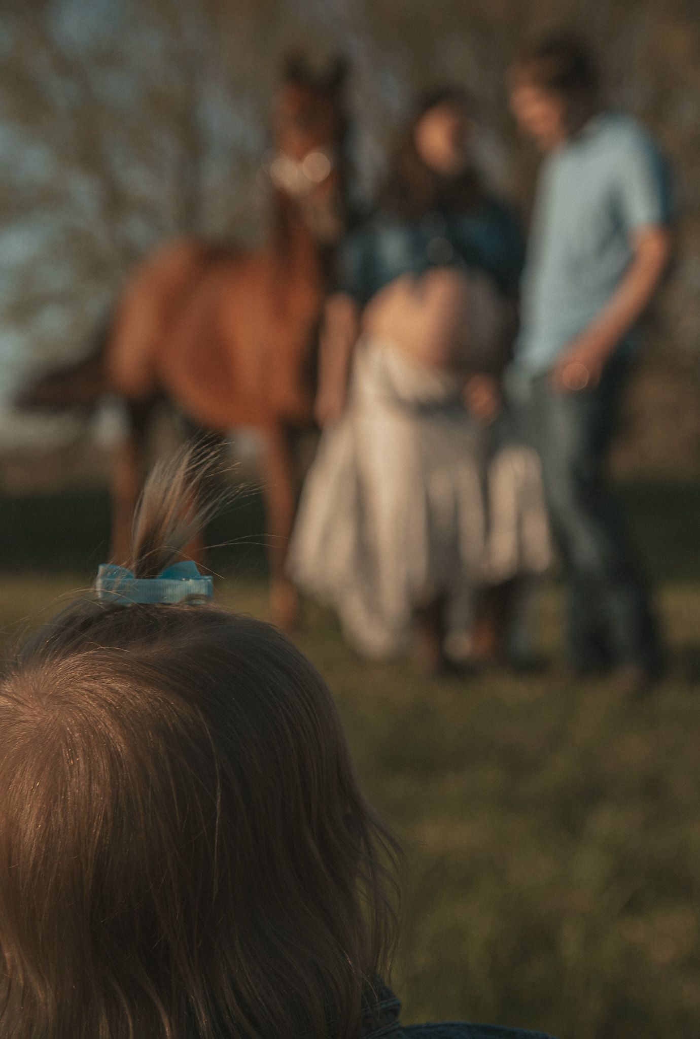 A child with a hair clip watches a couple and their horse in a blurred outdoor maternity photo session.