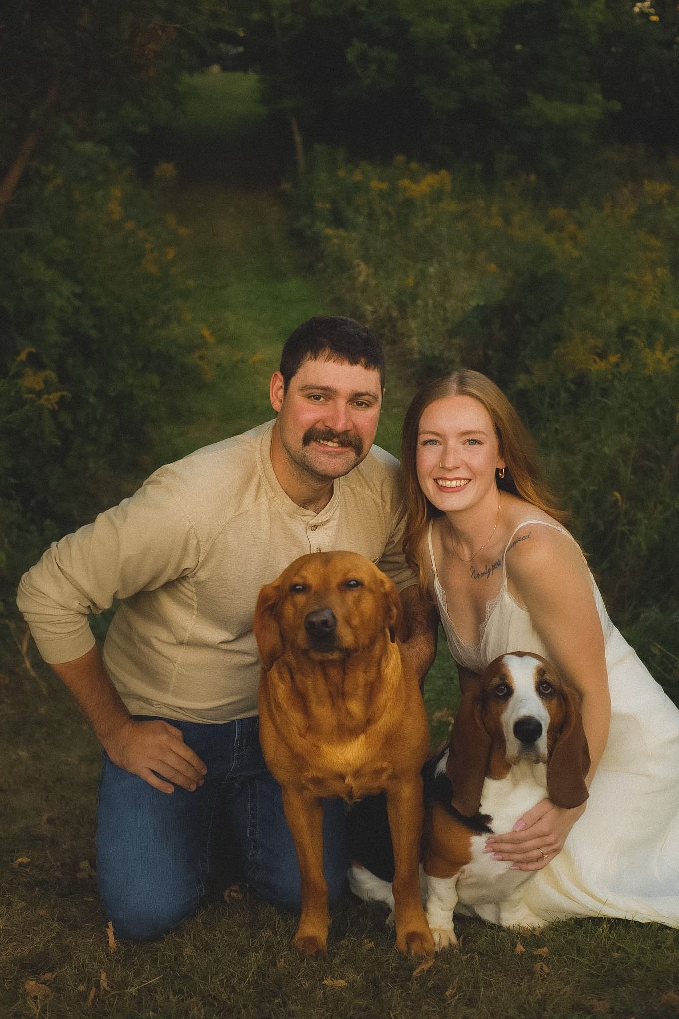 A smiling couple poses outdoors with a golden Labrador and a Basset Hound in a lush green garden setting.