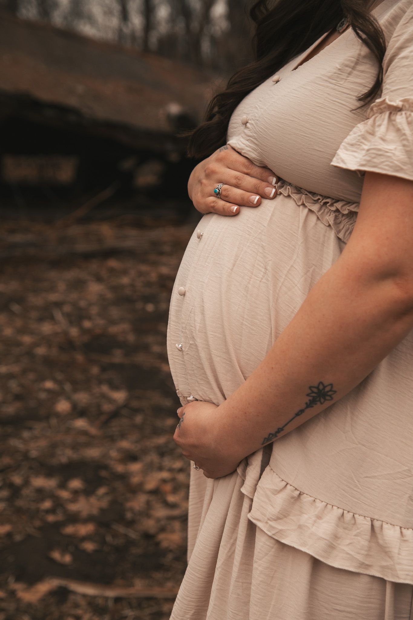 Pregnant woman in beige dress cradling baby bump outdoors, showing tattoo and turquoise ring.