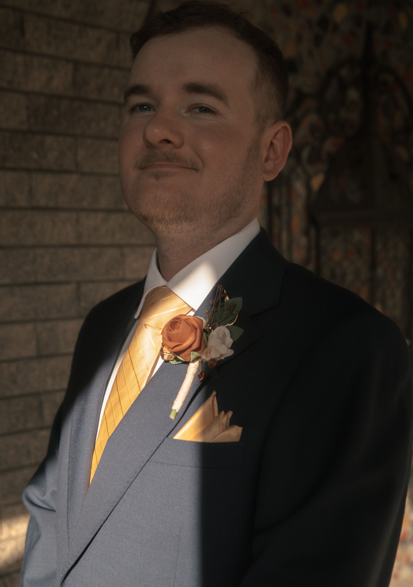 A person in a gray suit with a yellow tie and boutonniere poses for a formal portrait series against a brick wall.