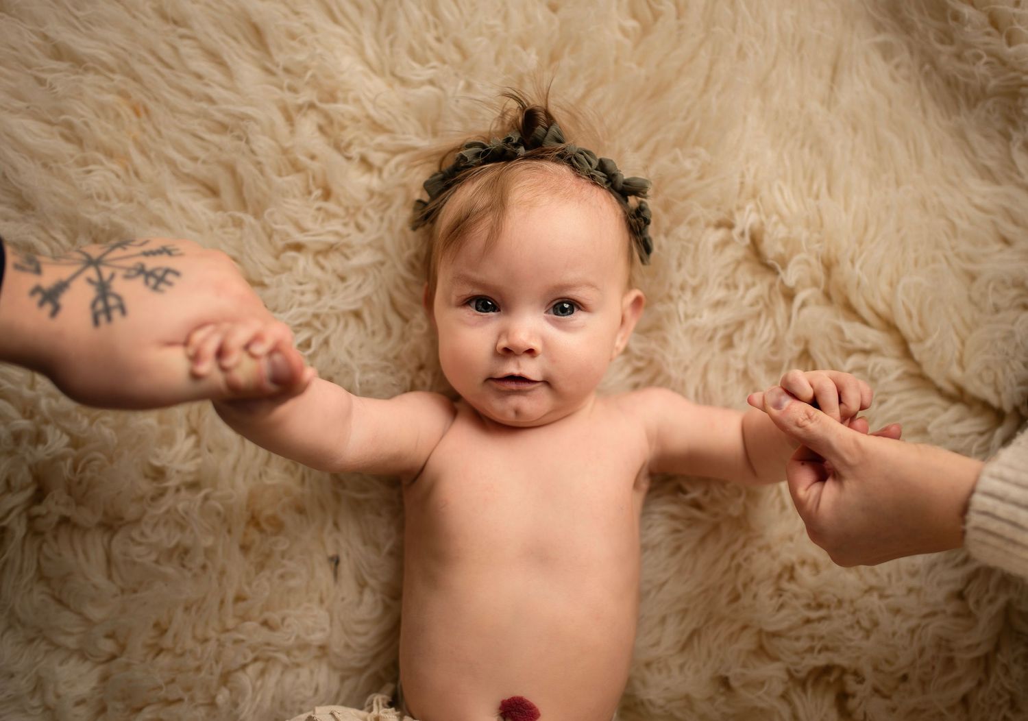Baby lying on beige fur rug with hands held by adults on either side.
