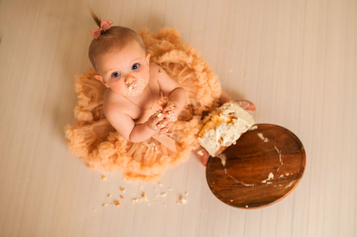 Overhead view of baby in peach tulle surrounded by cake smash mess on wooden floor.
