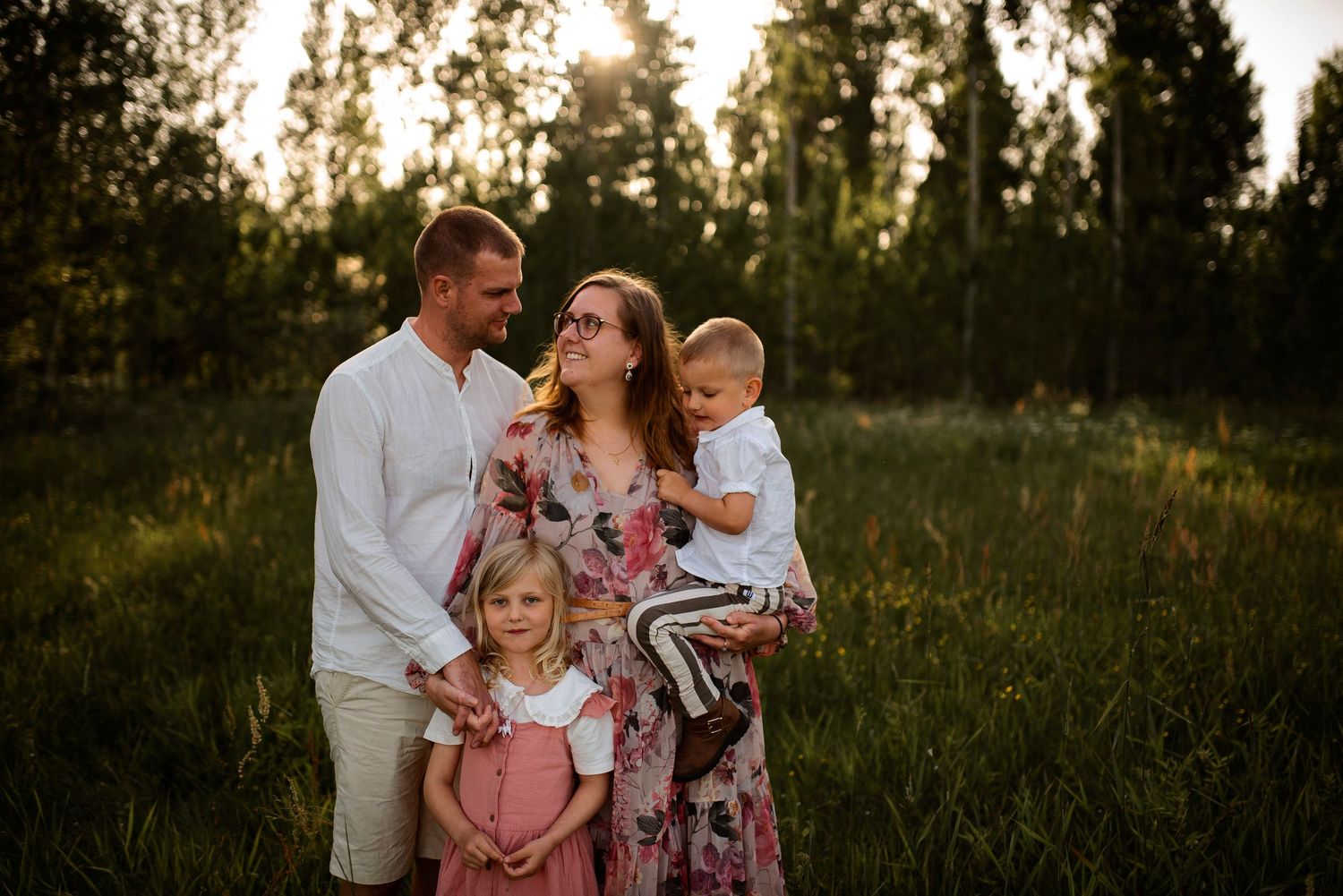 Family stands together in a field with sun setting through trees behind them.