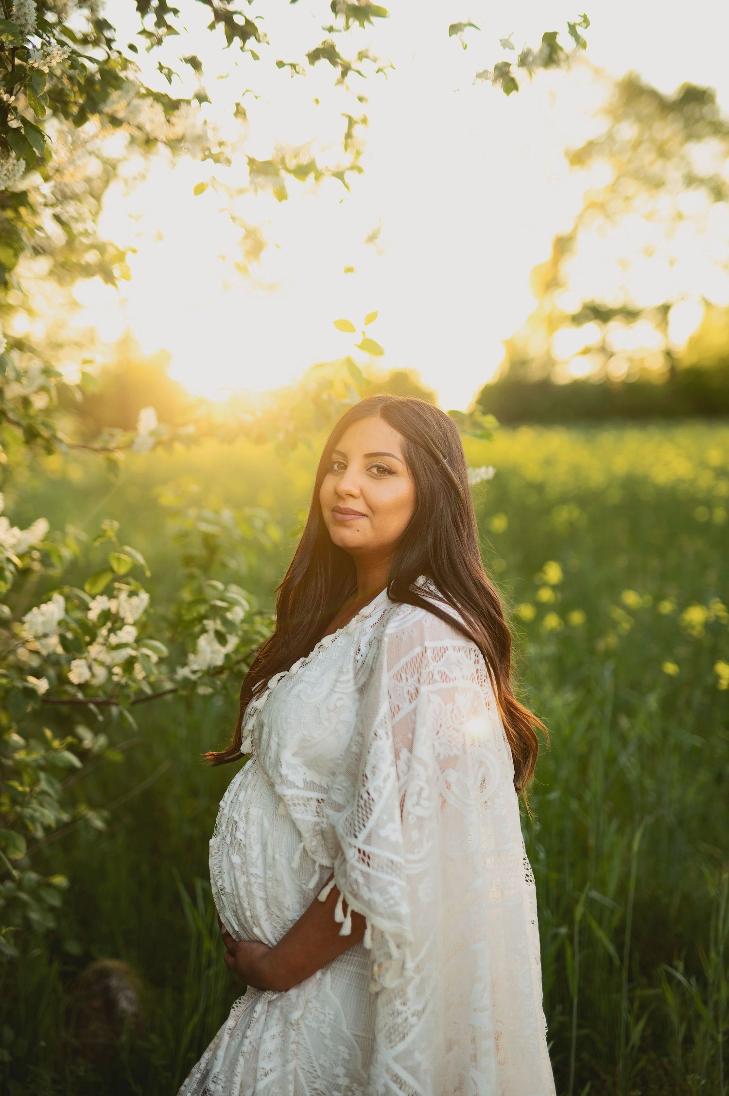 Expectant mother in white lace dress poses in flowering meadow during golden sunset.