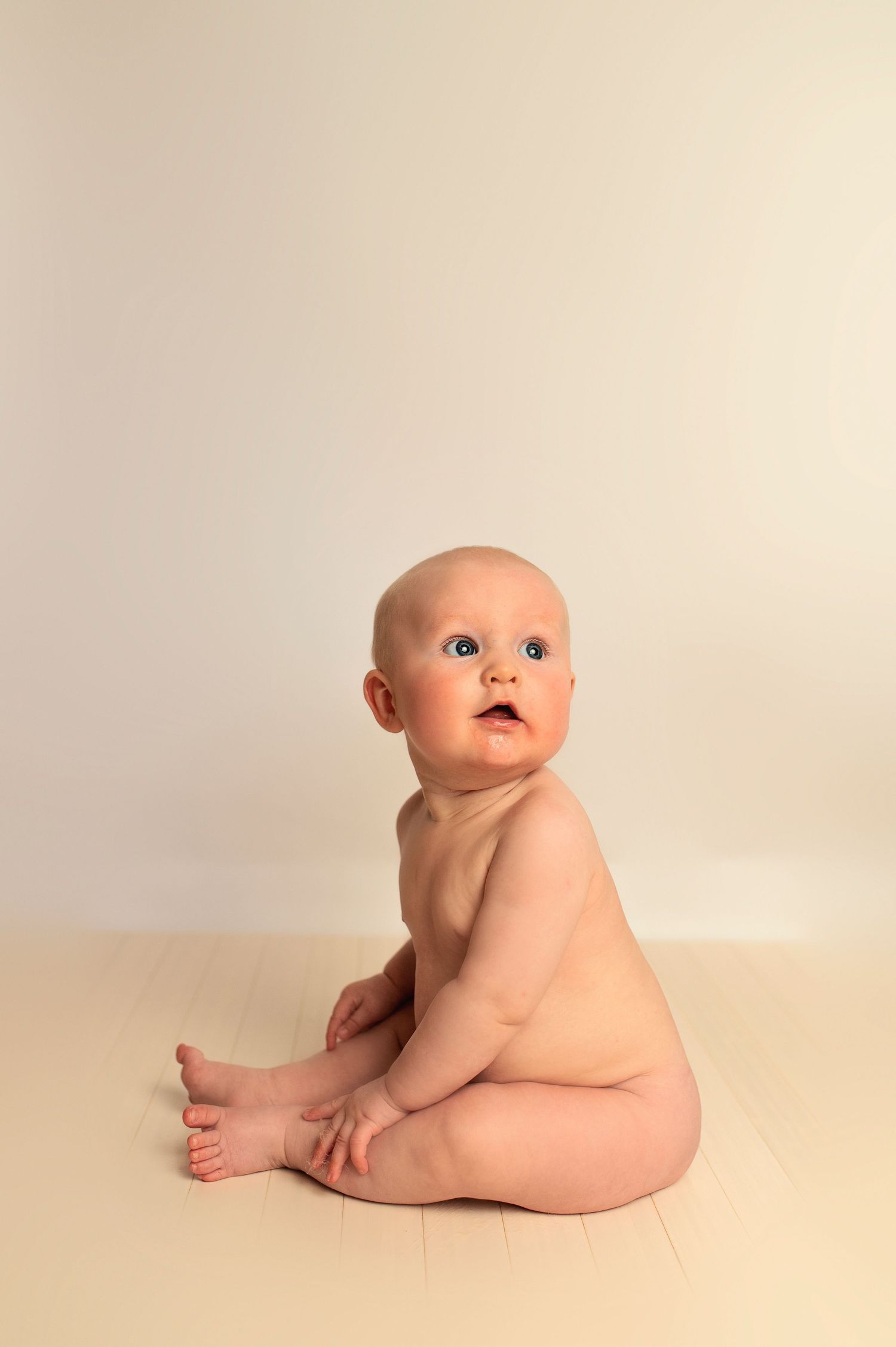 Baby sitting on beige backdrop looking up with curious expression.