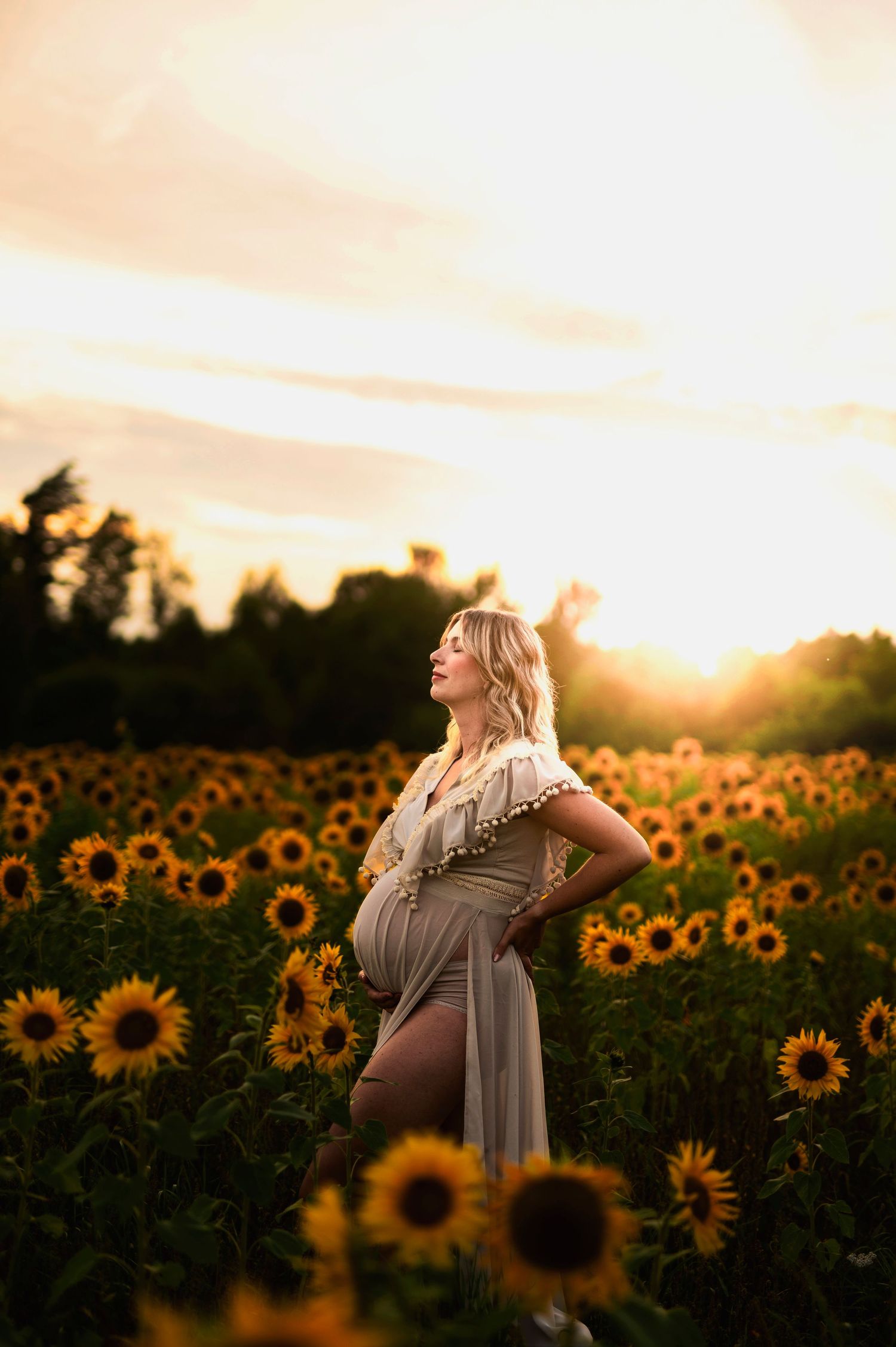 Maternity photo in flowing dress standing among blooming sunflowers at golden sunset.
