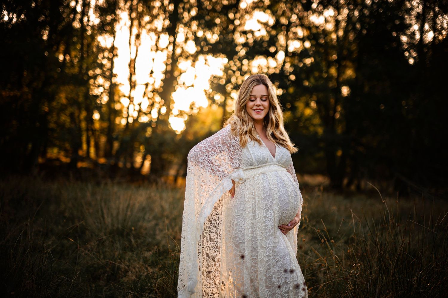 Maternity portrait in lace dress standing in sunlit field at golden hour.