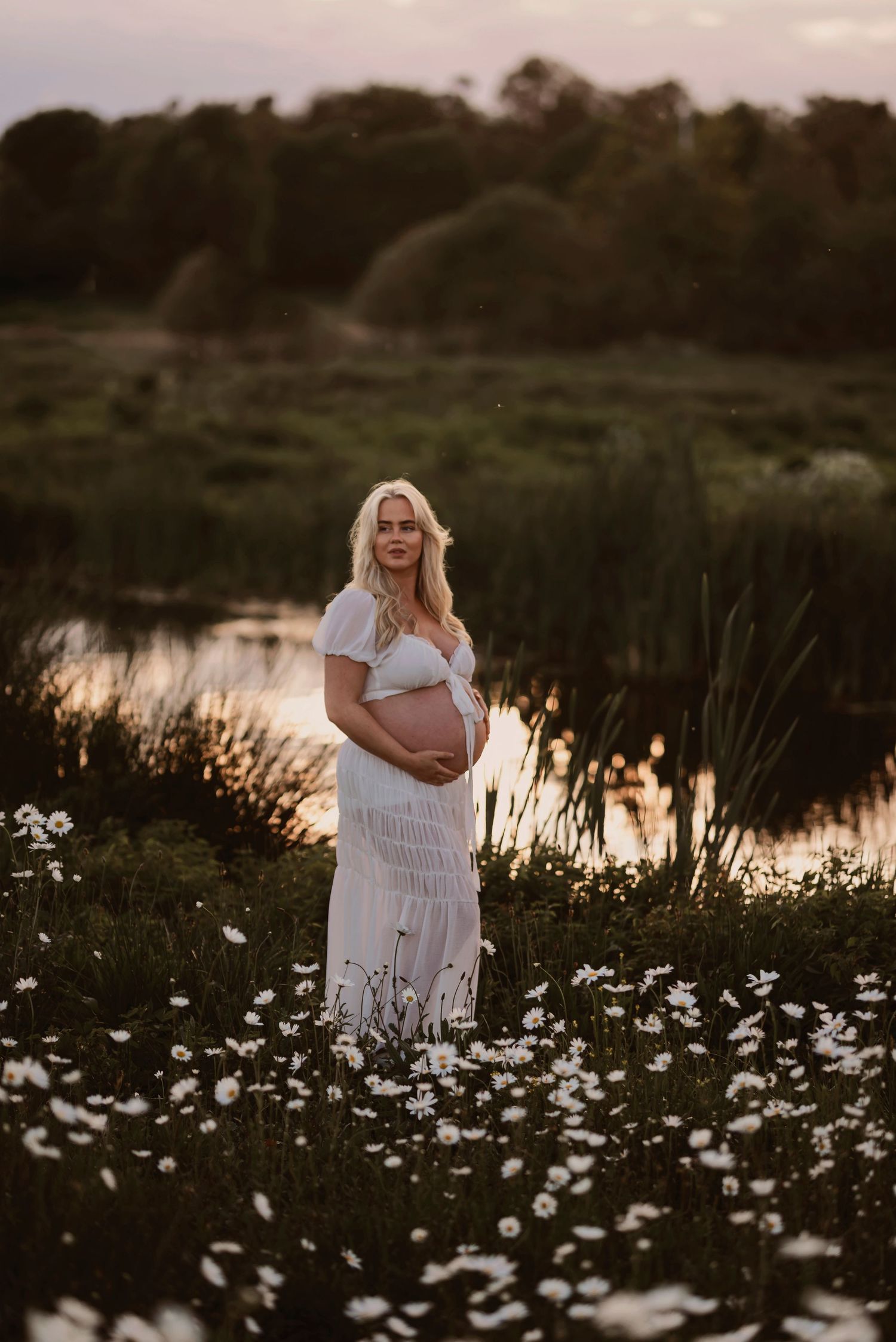 Person in a white dress stands among blooming white daisies near a tranquil pond at sunset.