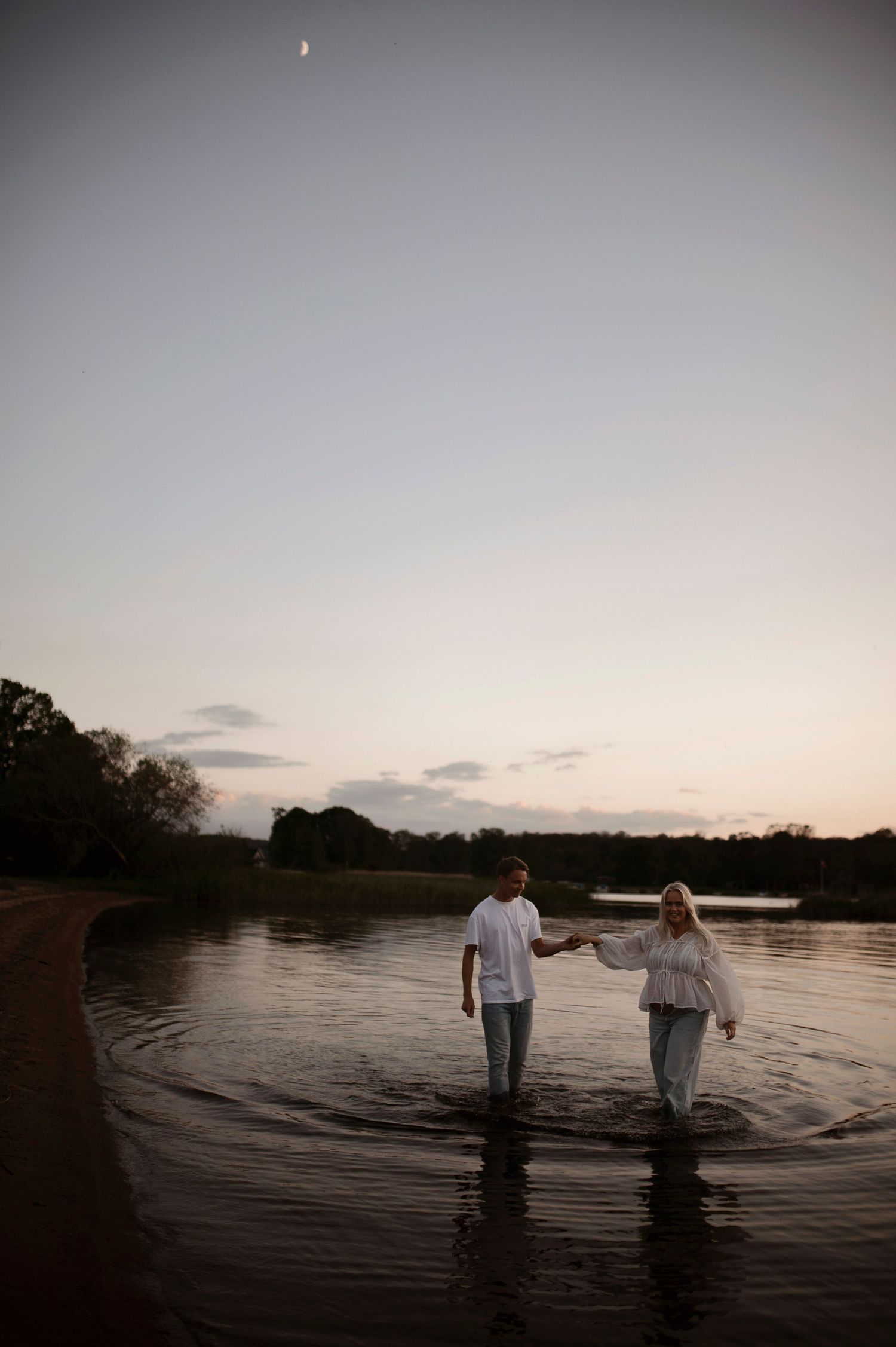 A couple holds hands while standing in shallow water during sunset.