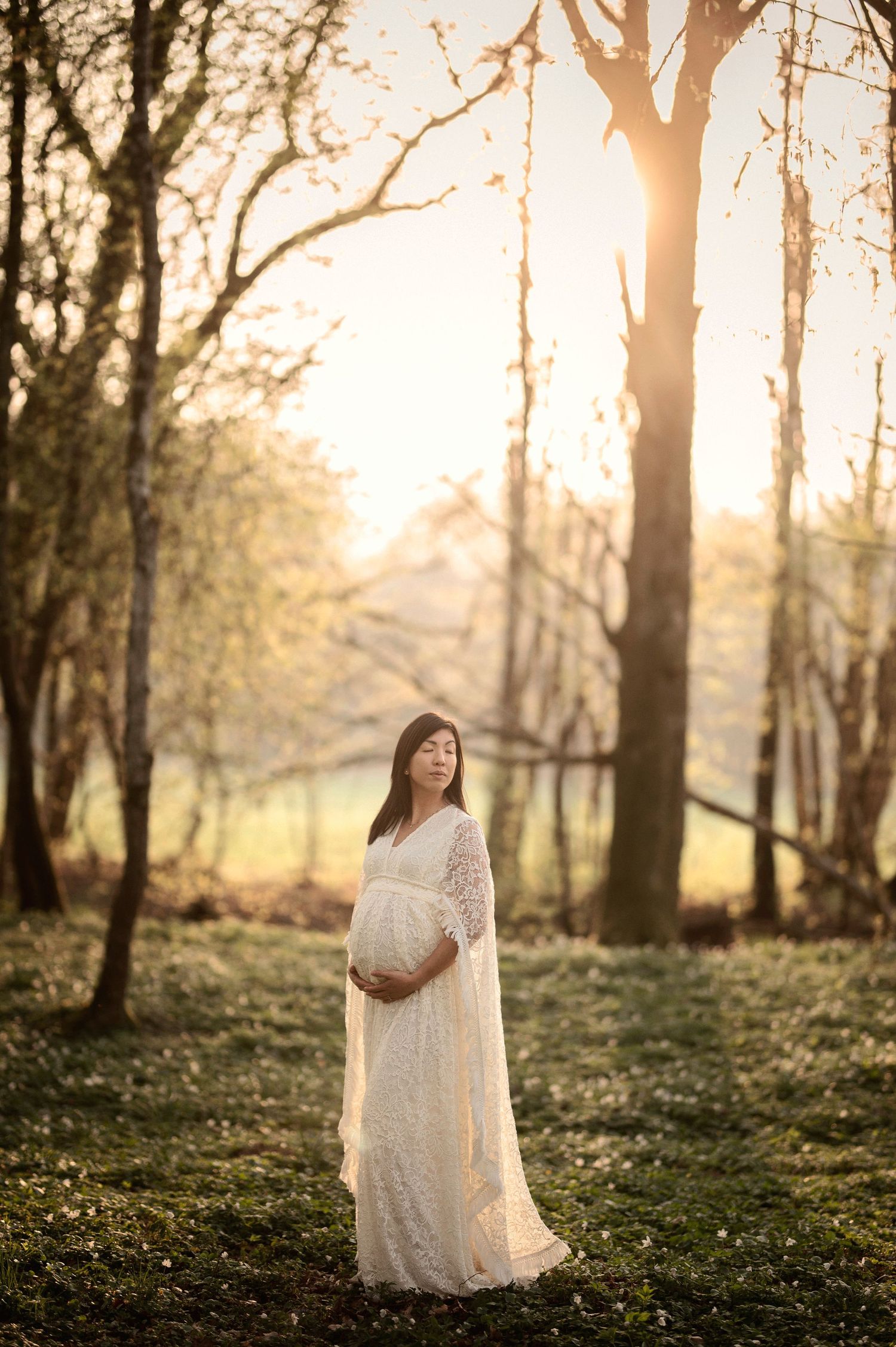 Ethereal maternity photoshoot in a sunlit forest with white lace dress and golden hour lighting.