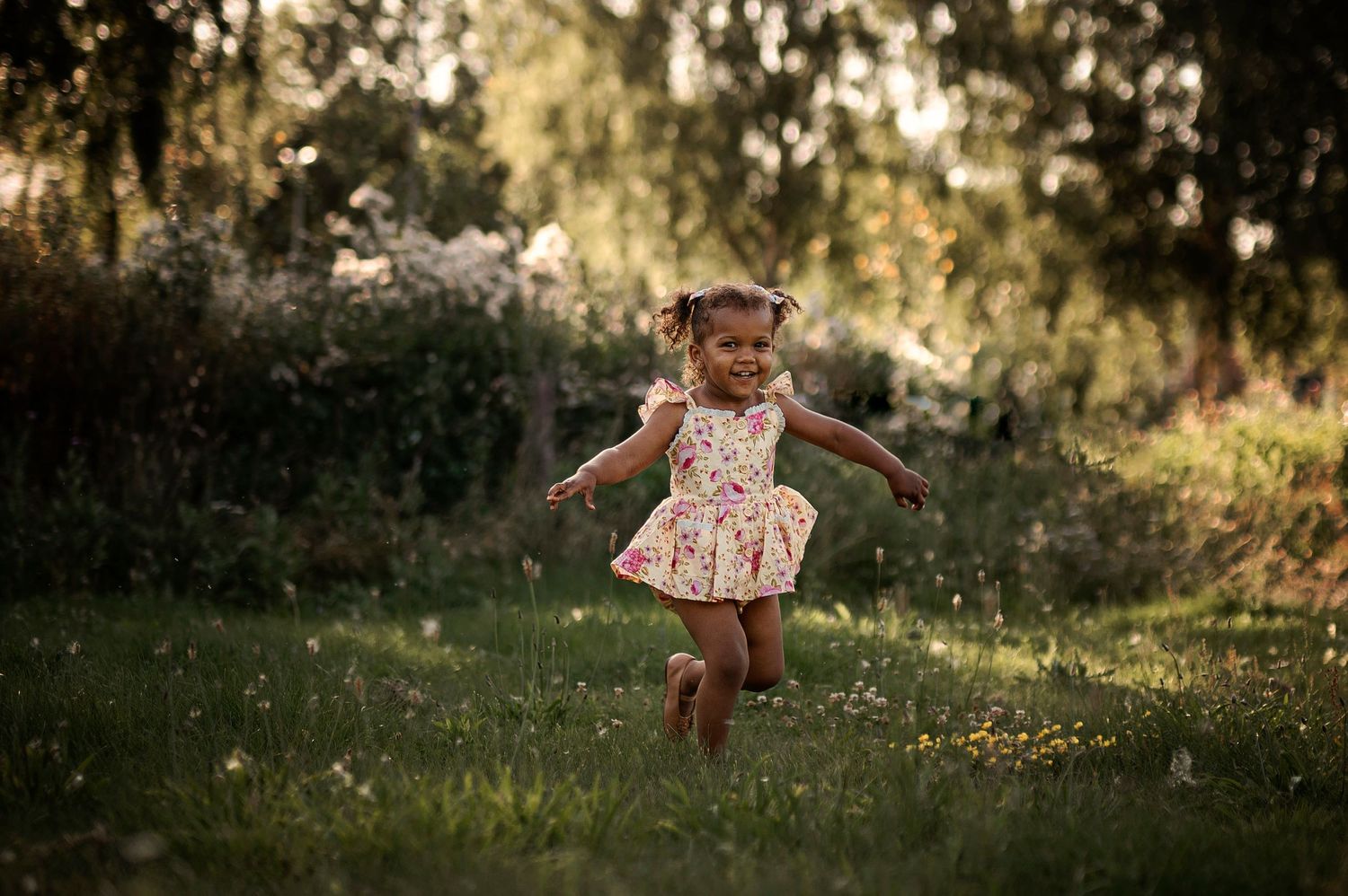 A young child in a floral dress jumps playfully in a sunlit garden.