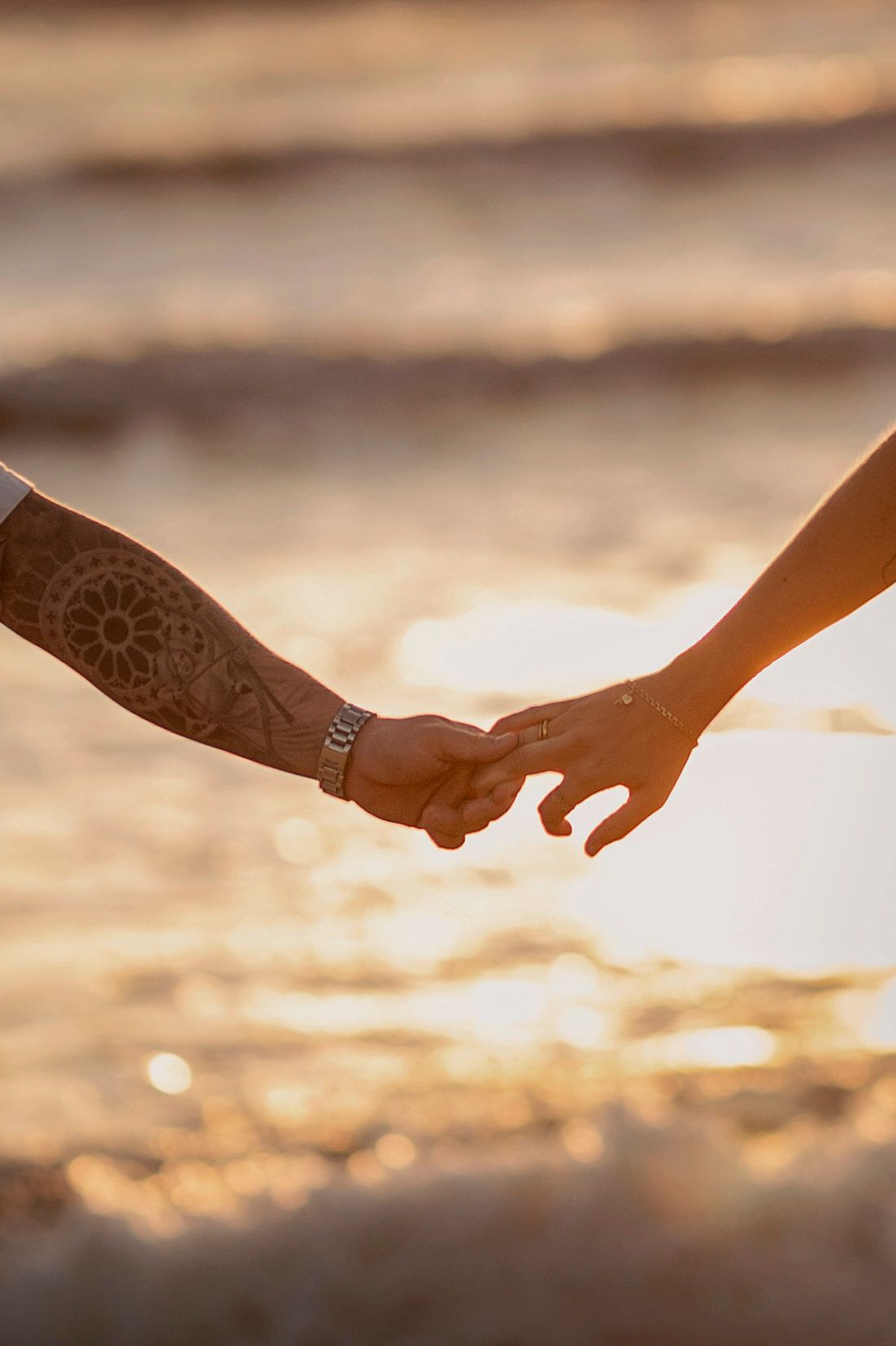 Two people holding hands on a beach at sunset with one person showing an intricate henna tattoo design on their arm.
