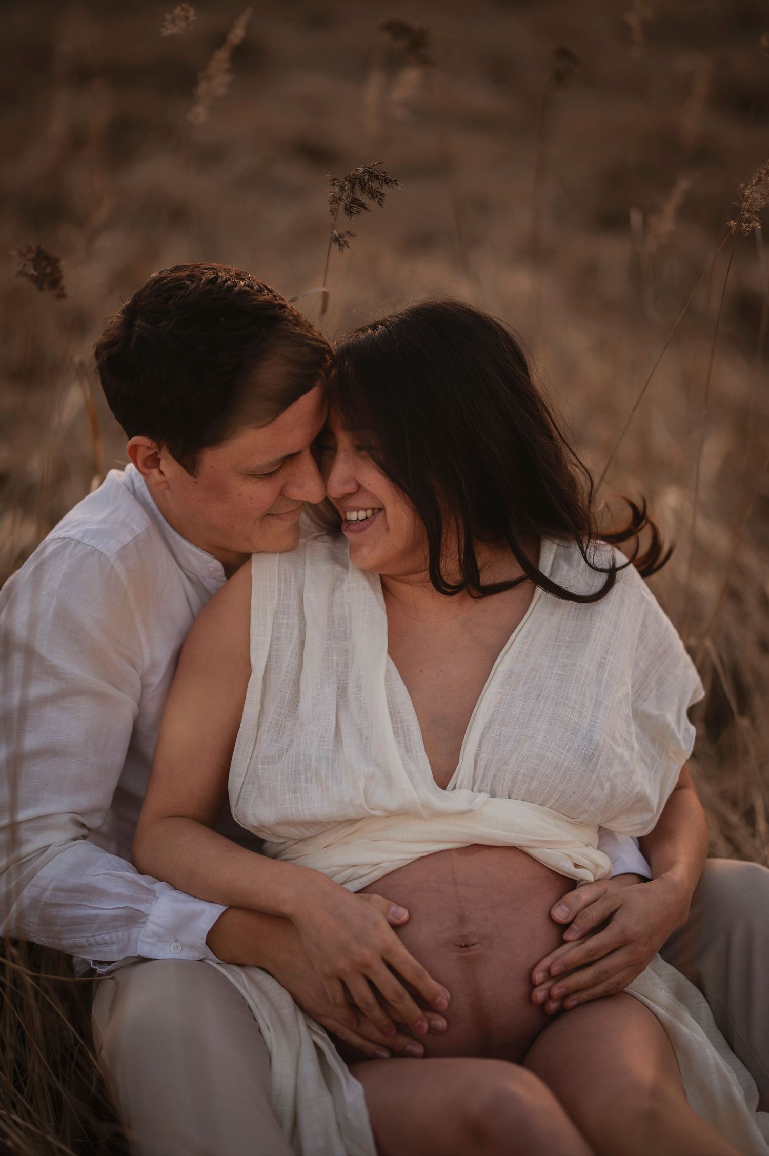 A tender maternity photo shows a couple in white clothing embracing in a sun-lit field while touching the pregnant belly.